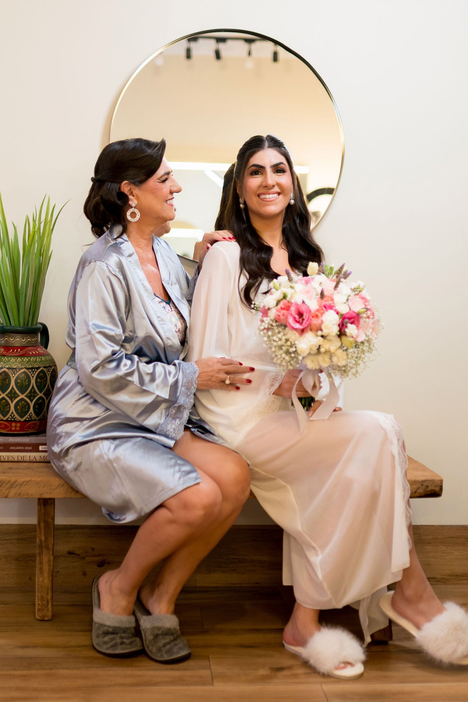Two women in satin robes sit on a bench, one holding a pink bouquet, smiling before a round mirror.