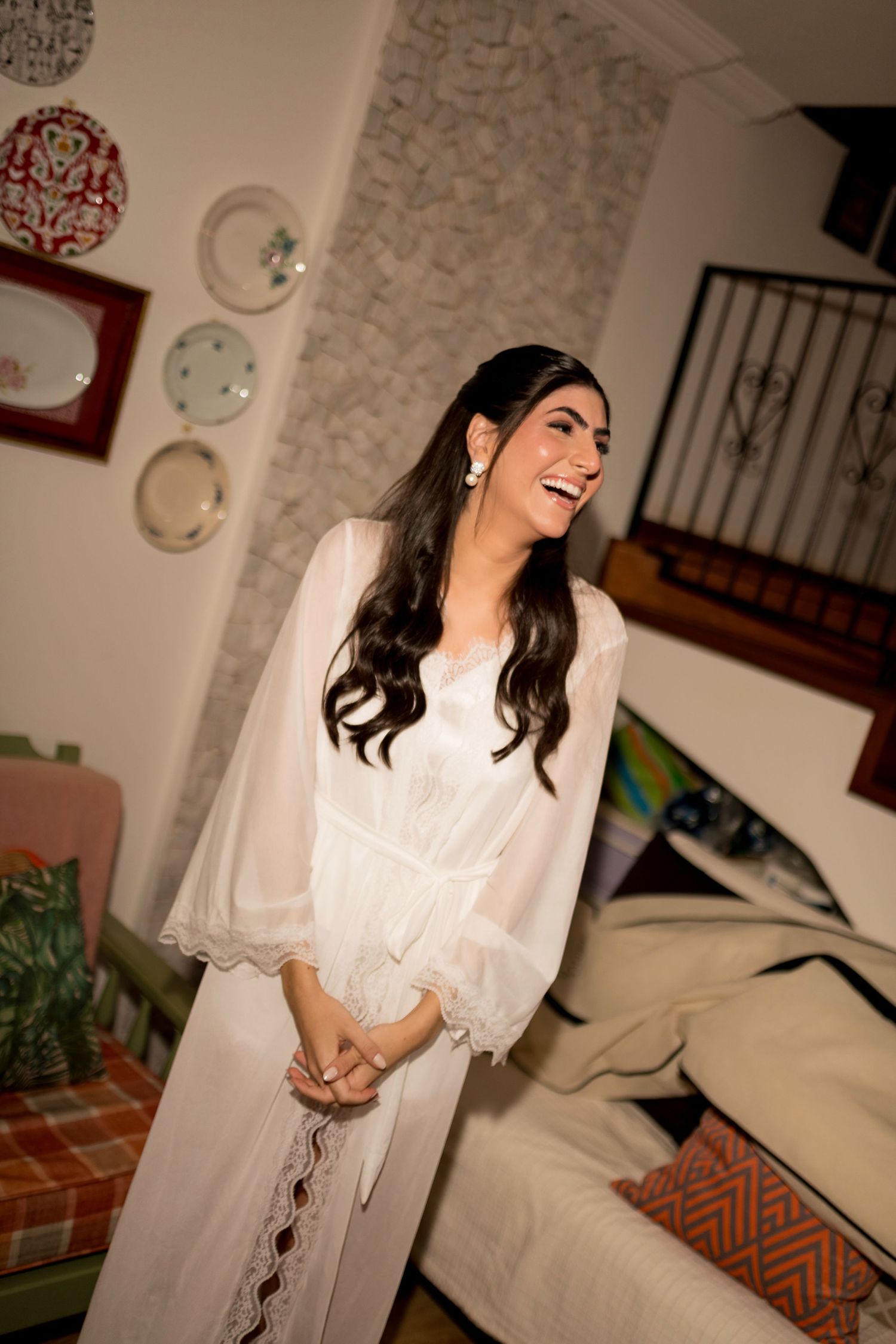 A smiling woman in a white lace dress with billowy sleeves stands in a cozy decorated room.