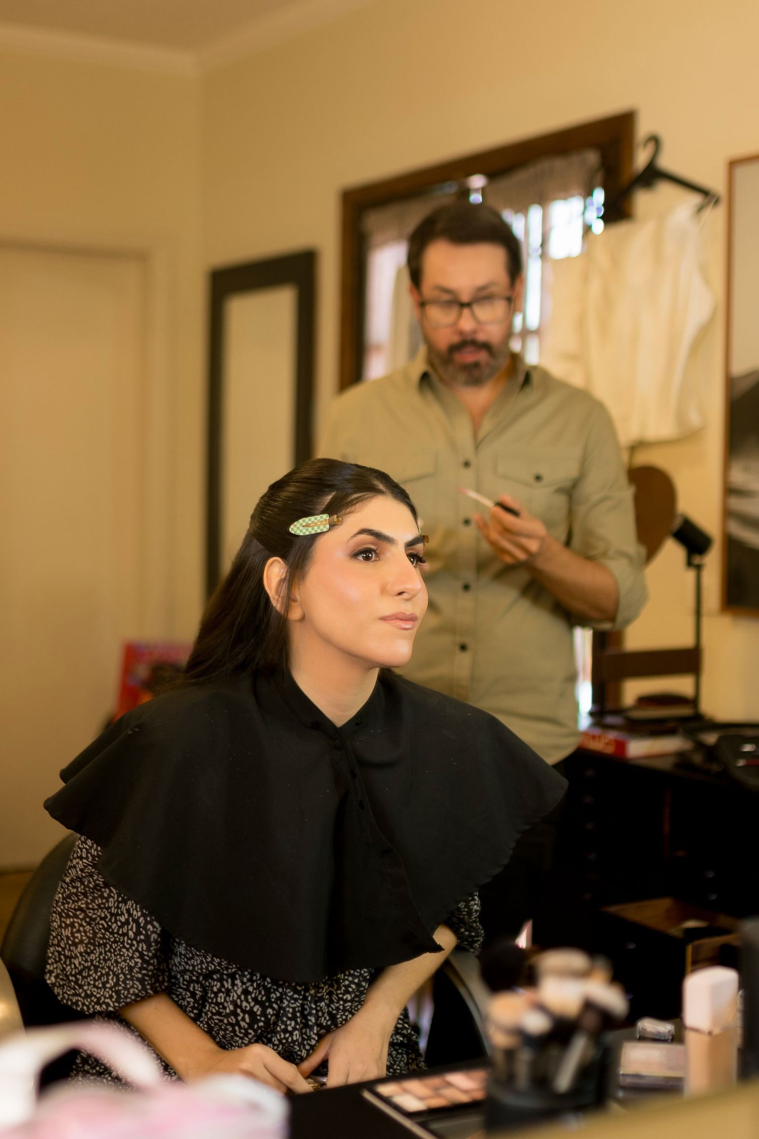A woman in a black cape sits at a makeup vanity while a bearded male makeup artist works behind her.