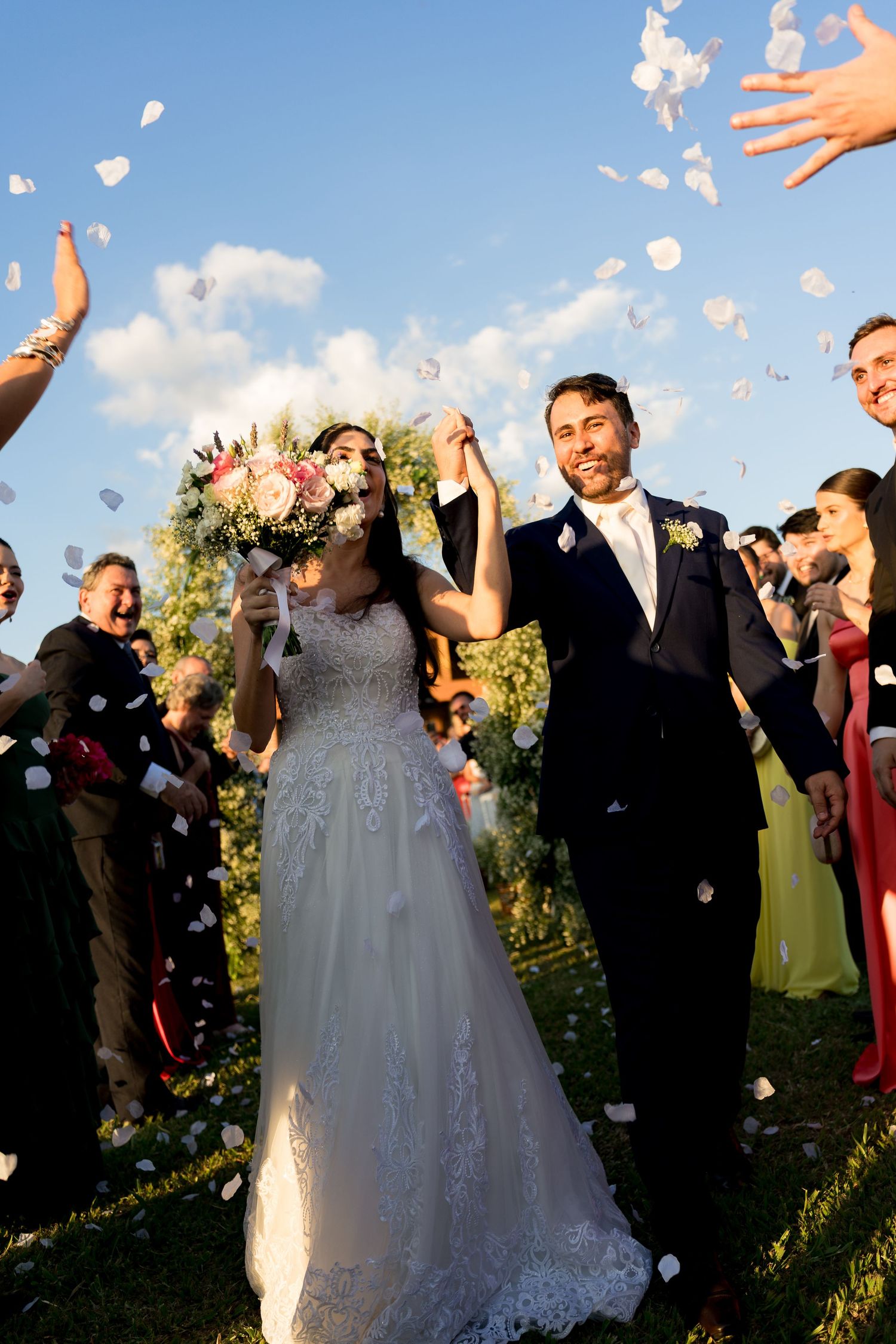 Joyful newlyweds walk through flower petal confetti shower, bride holding bouquet, guests celebrating outdoors under blue sky.