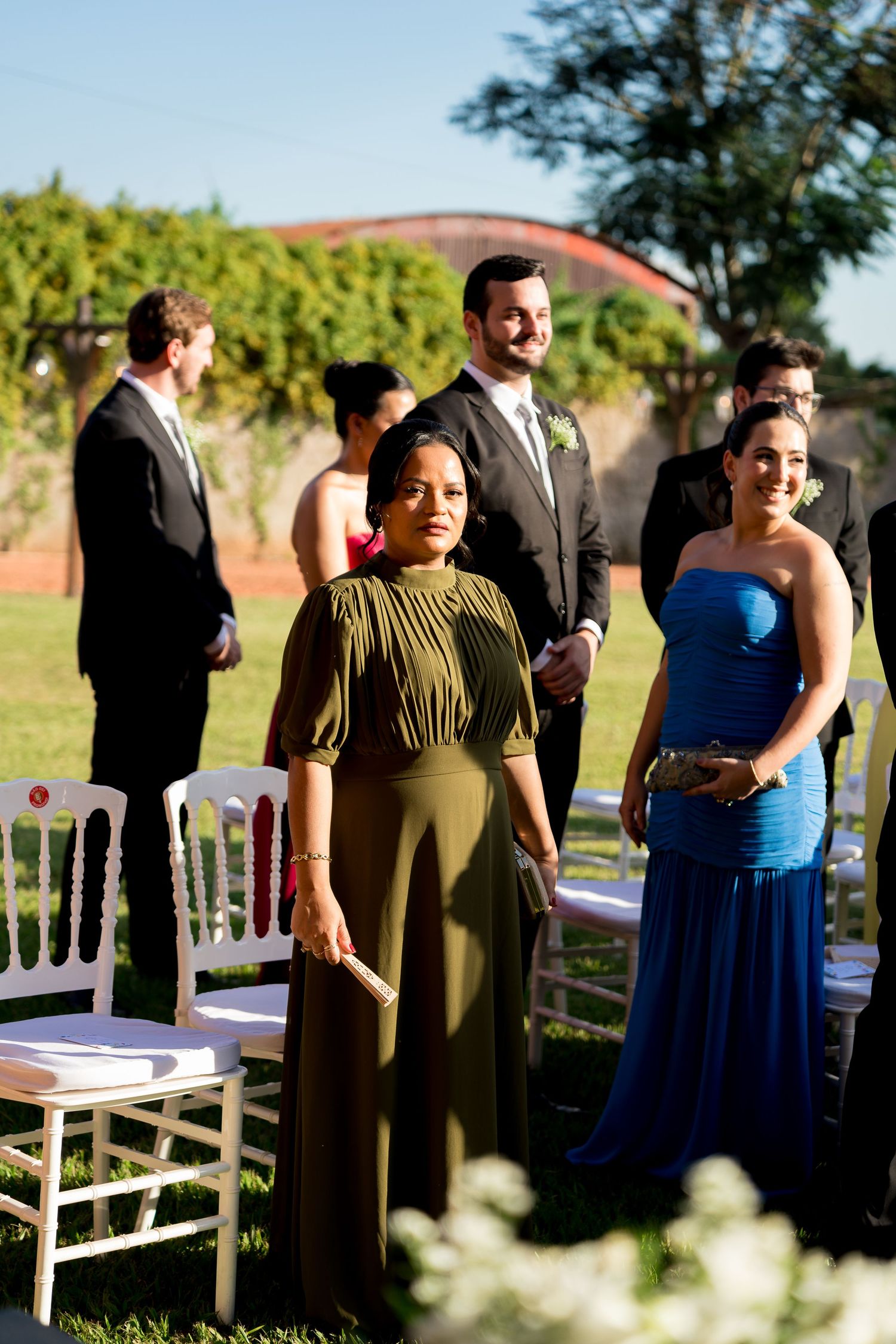 Two elegantly dressed women stand at an outdoor wedding ceremony, one in olive green and one in a blue strapless gown.