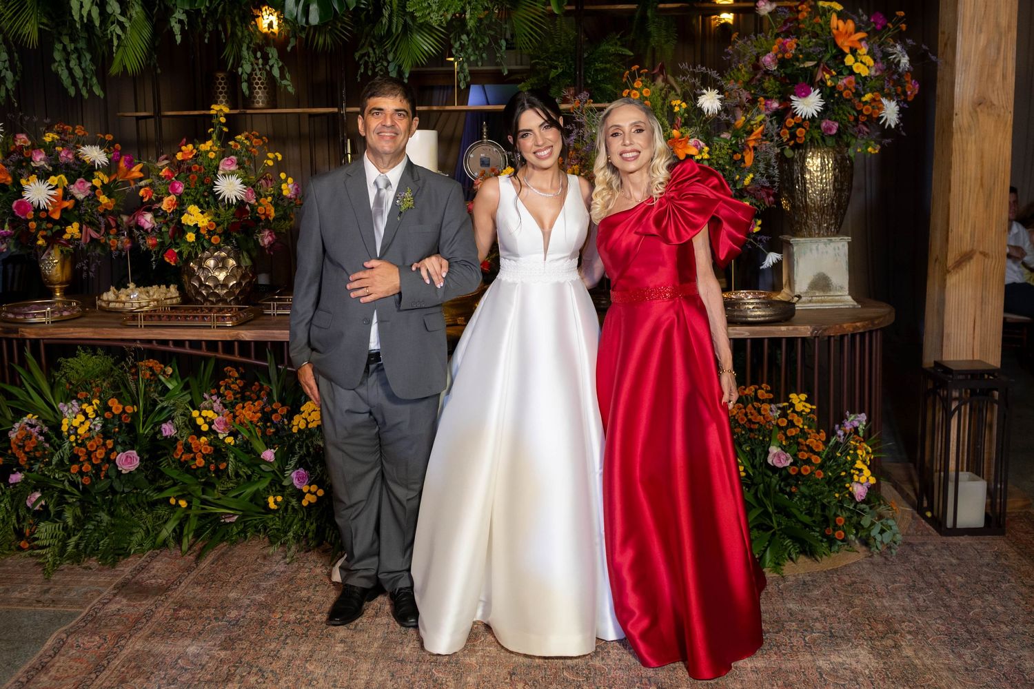 Bride in white gown poses with groom in gray suit and woman in red dress at floral wedding reception.