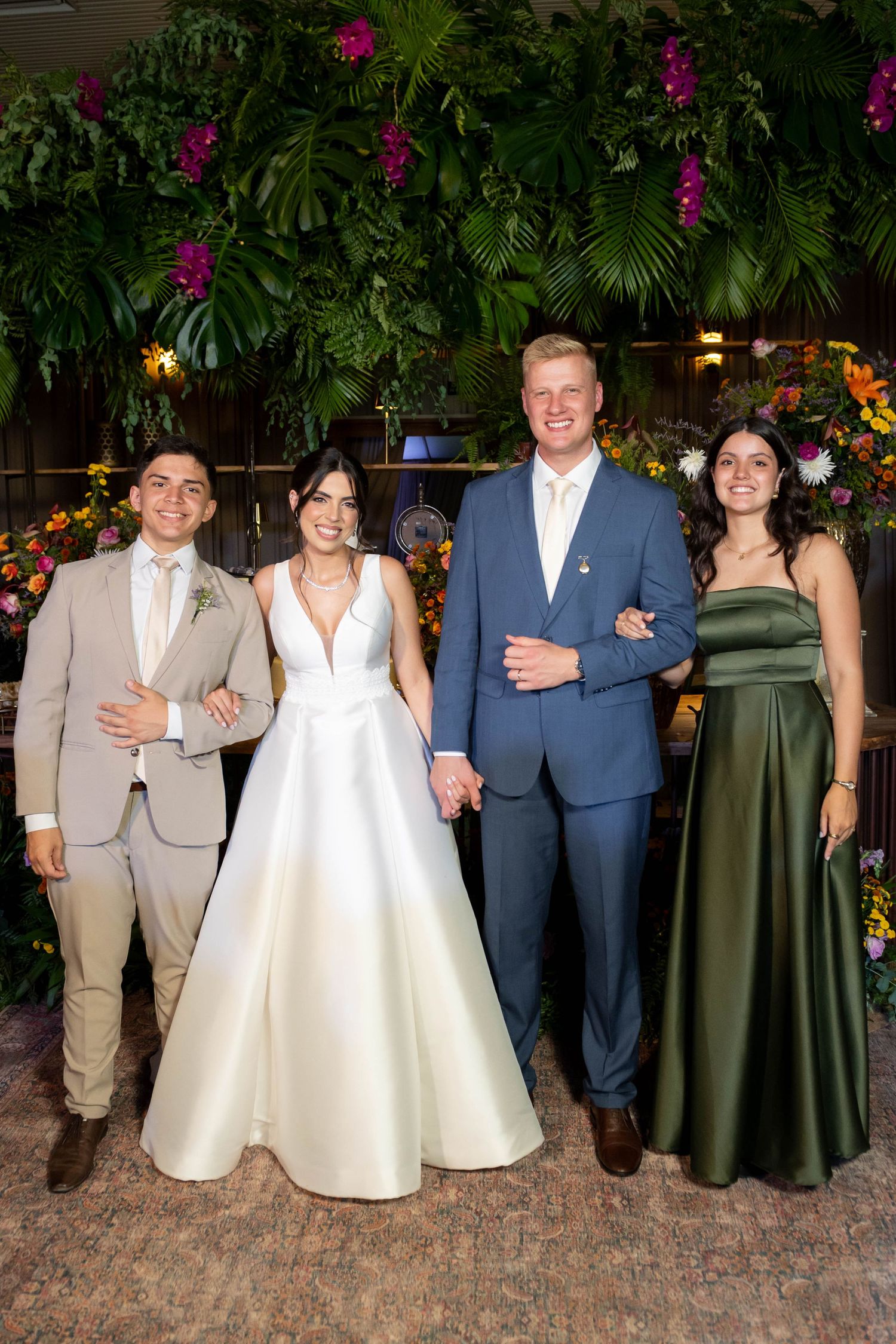 Bride and groom holding hands with two guests in front of a lush floral green wall backdrop at a wedding reception.