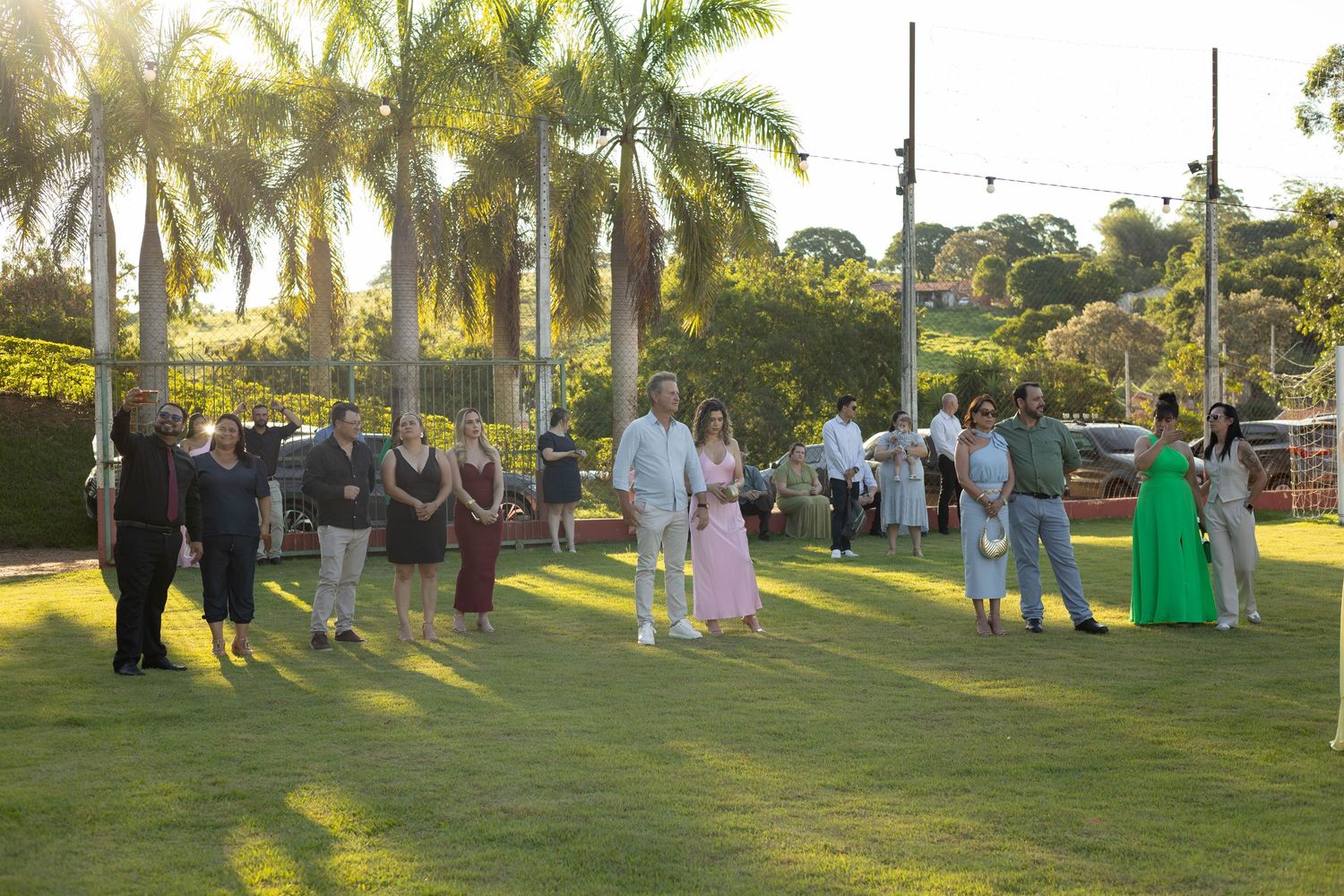 Elegantly dressed guests gathered on lush green lawn surrounded by tall palm trees at an outdoor evening event.