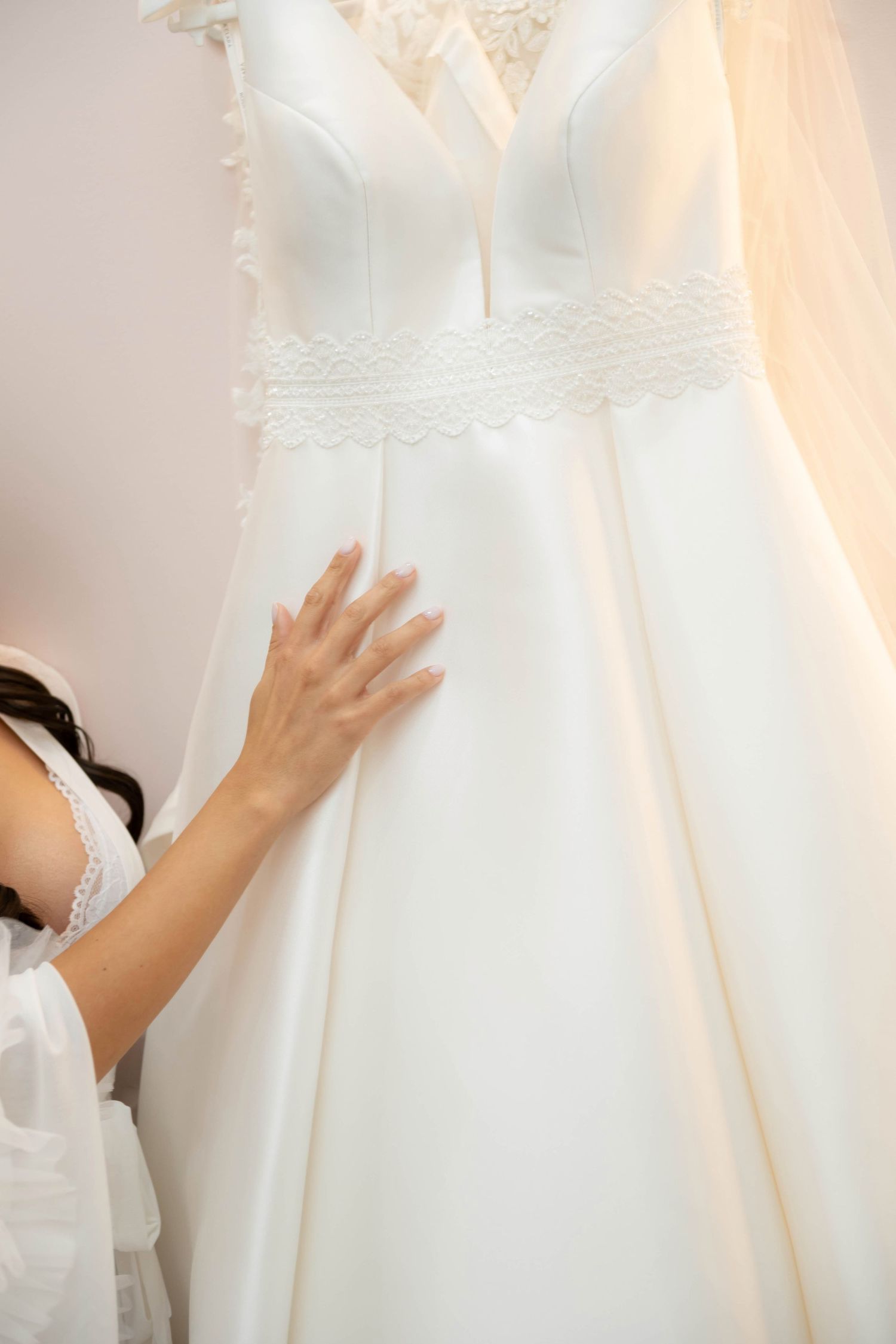 Bride gently touching an elegant white wedding dress with lace details hanging on display in a bridal boutique.