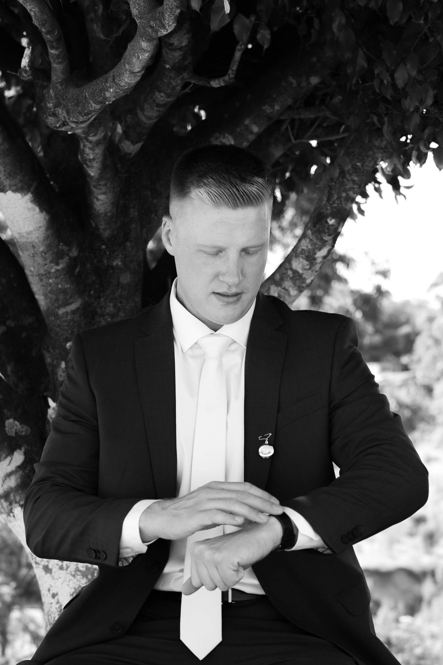 Black and white portrait of a young man in a suit and tie, seated under a tree, looking down thoughtfully.