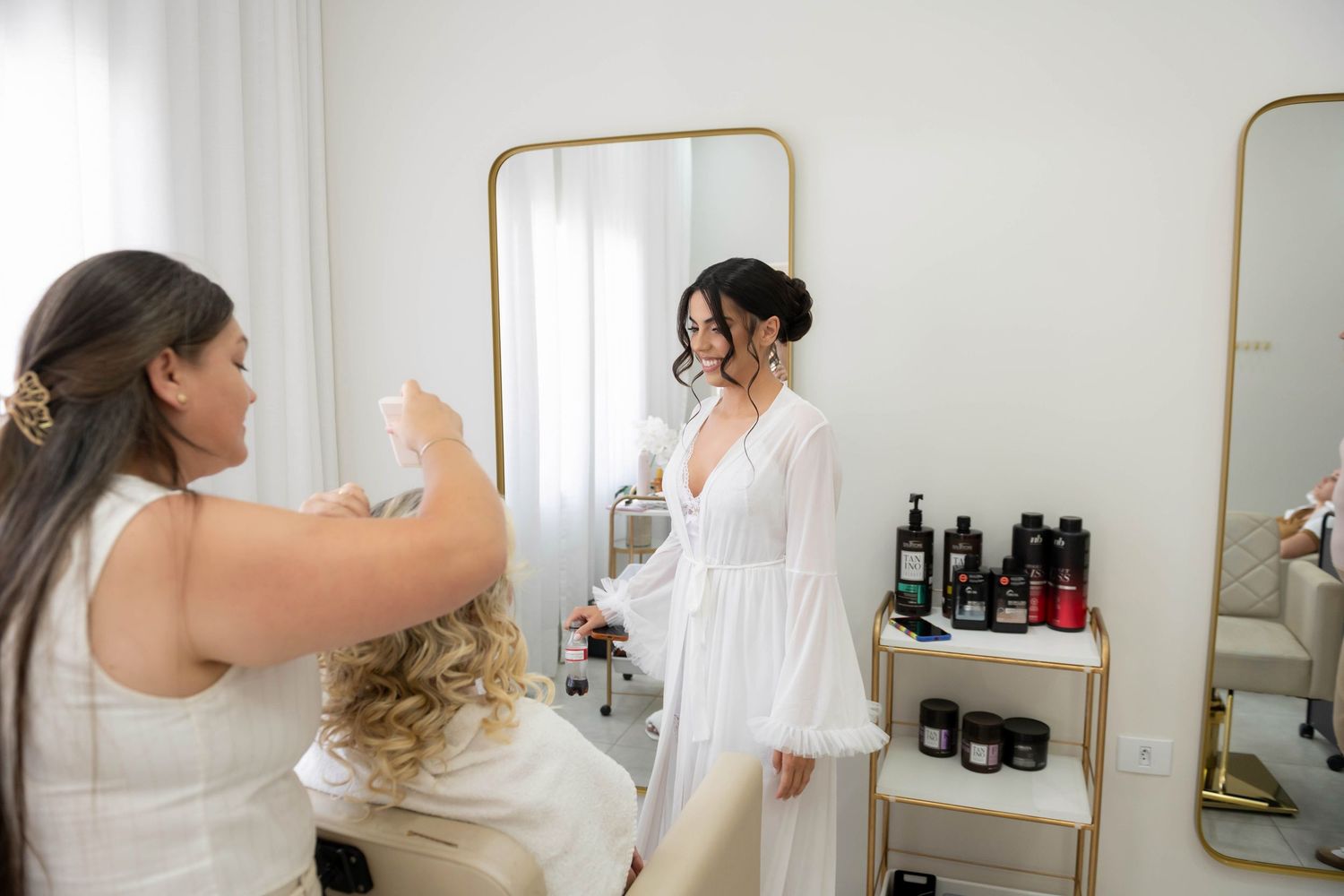 Bride in white robe smiles at stylist during wedding morning preparations in bright salon with gold mirrors.