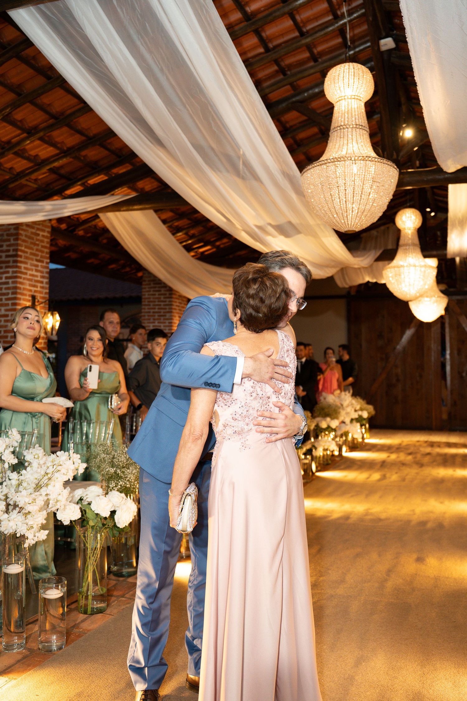 Groom in blue suit embracing woman in pink gown at elegant wedding venue with chandeliers and white floral arrangements.