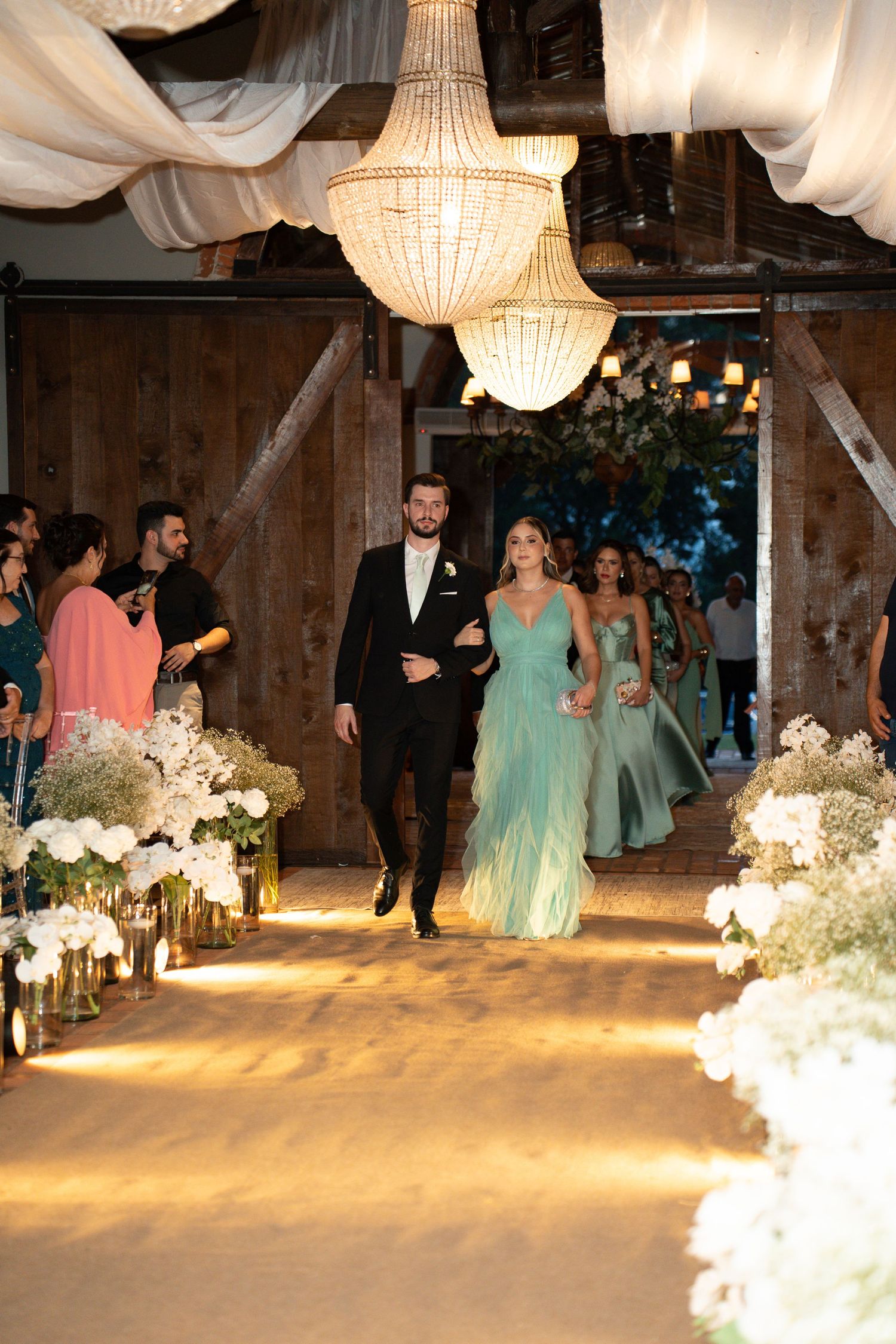 Couple walks down floral-lined wedding aisle in rustic barn venue with chandeliers and bridesmaids in teal gowns.
