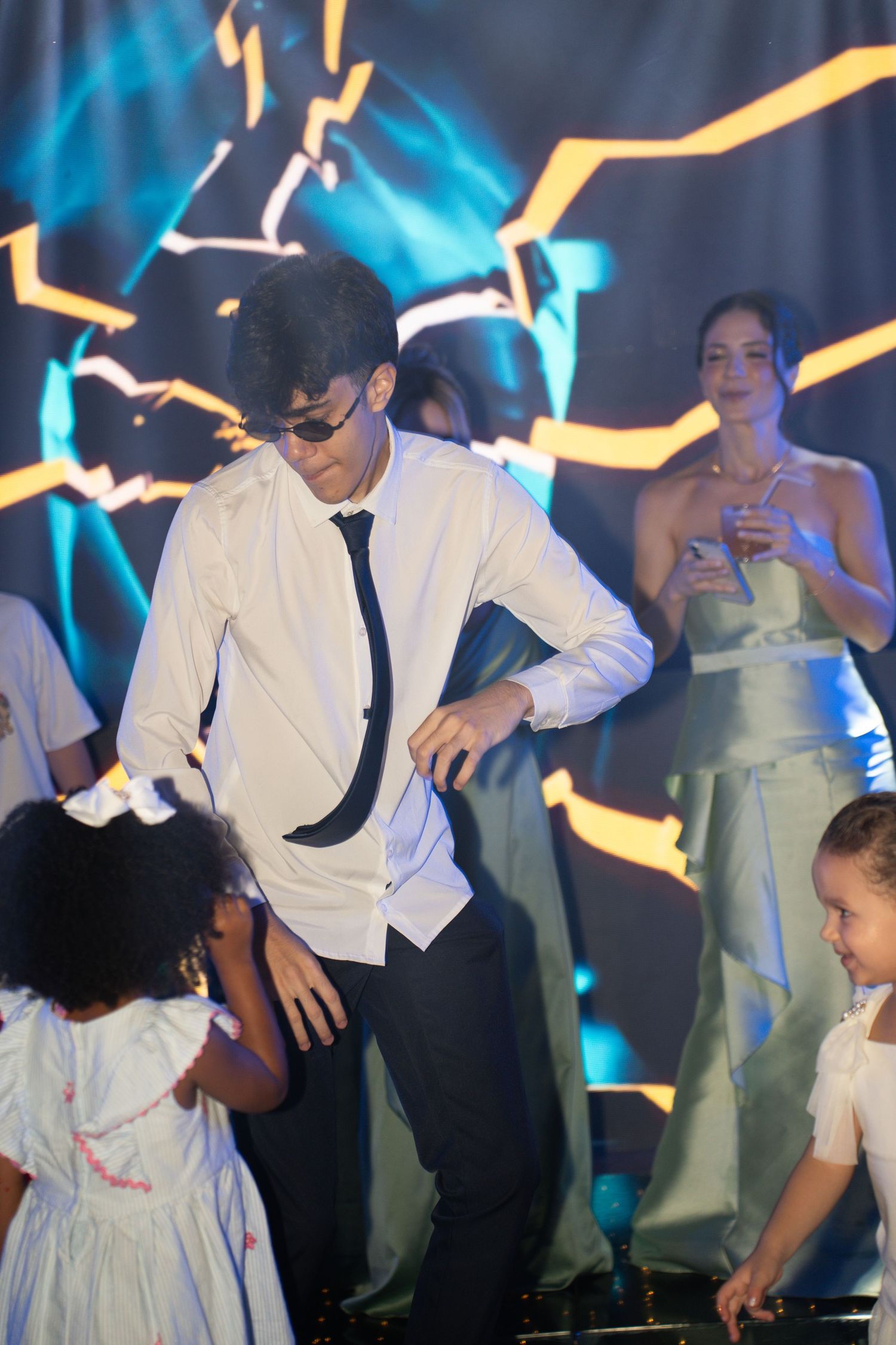 Man in white shirt and tie dancing with children at a colorful event with glowing stage backdrop.