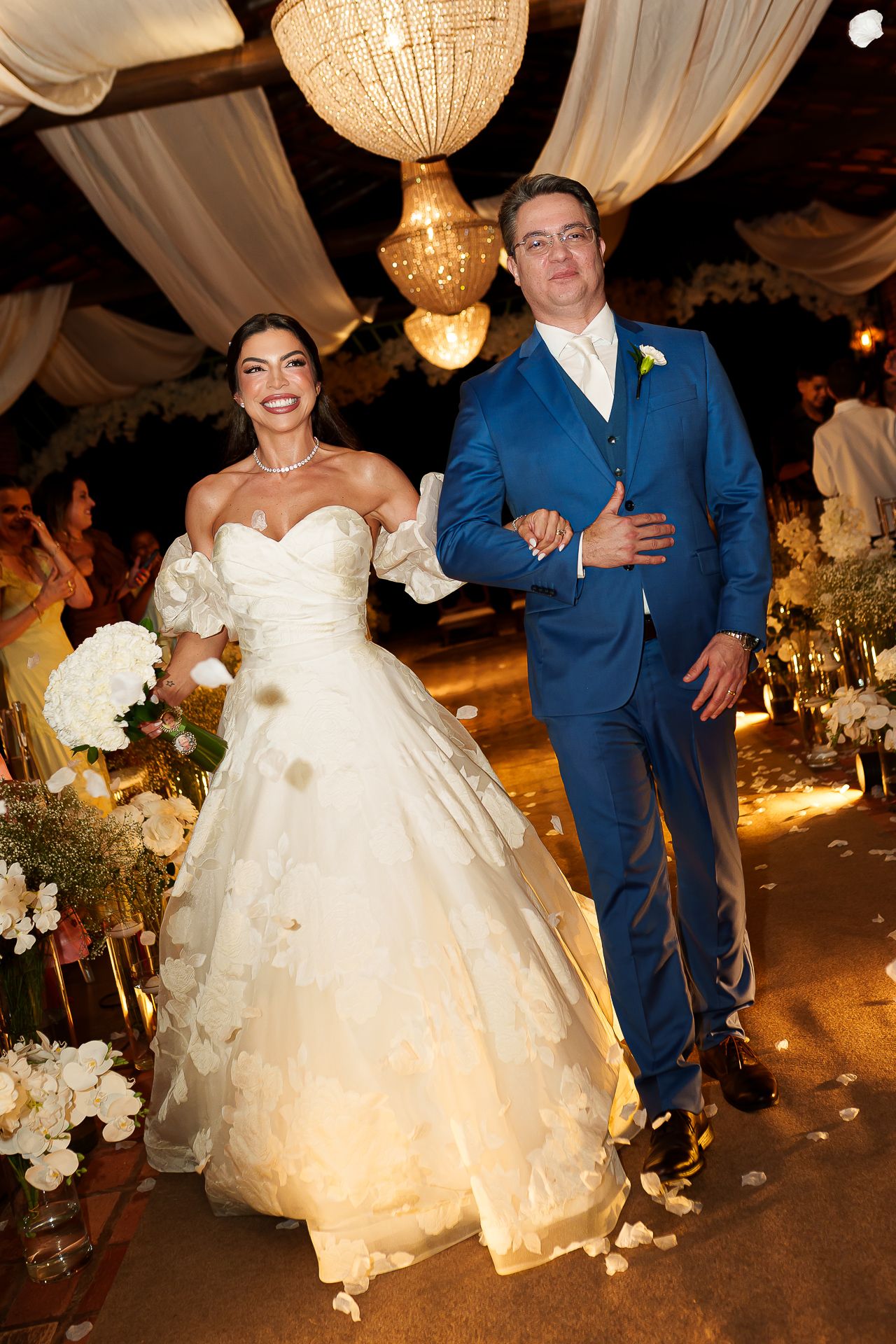 Bride in white ballgown and groom in blue suit walk together at elegant wedding reception with chandelier lighting.