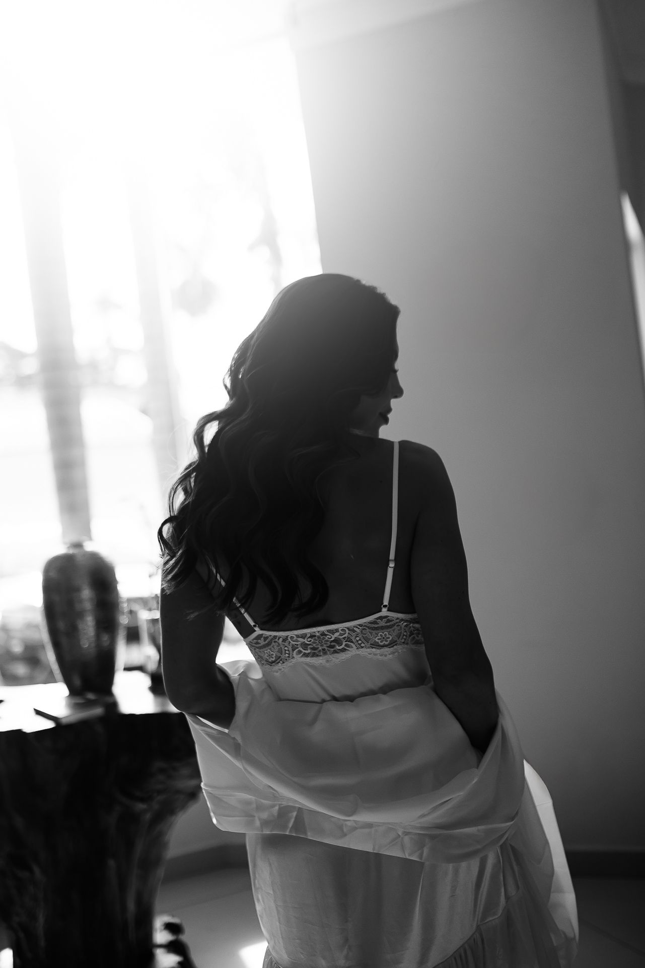 Black and white photo of a bride in white lace dress with long curly hair, standing near a bright window.