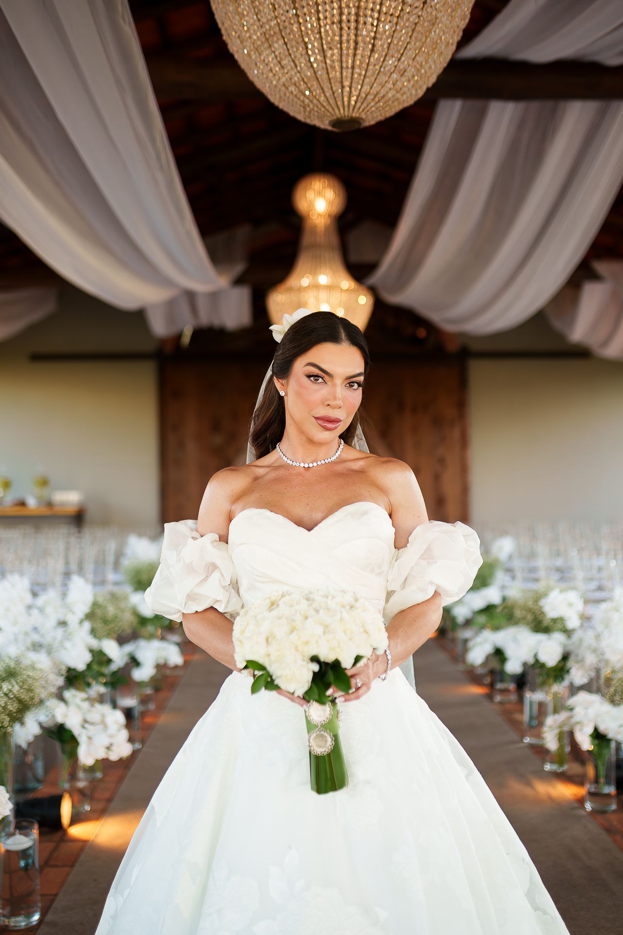Elegant bride in off-shoulder white ballgown holding white bouquet stands in decorated wedding venue with chandeliers.