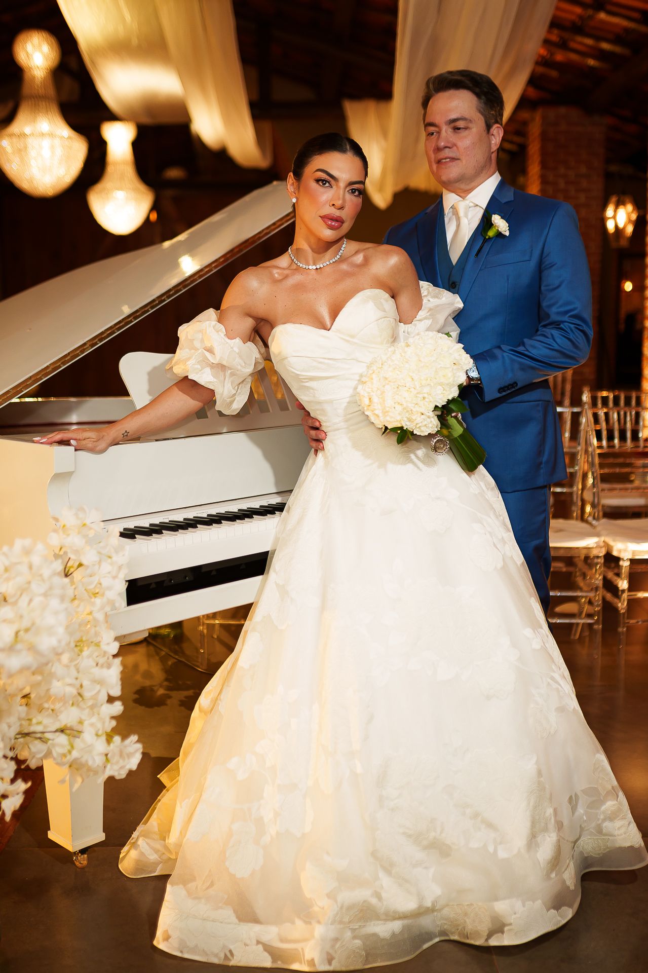 Bride in off-shoulder ball gown holds white bouquet beside groom in blue suit near white grand piano.