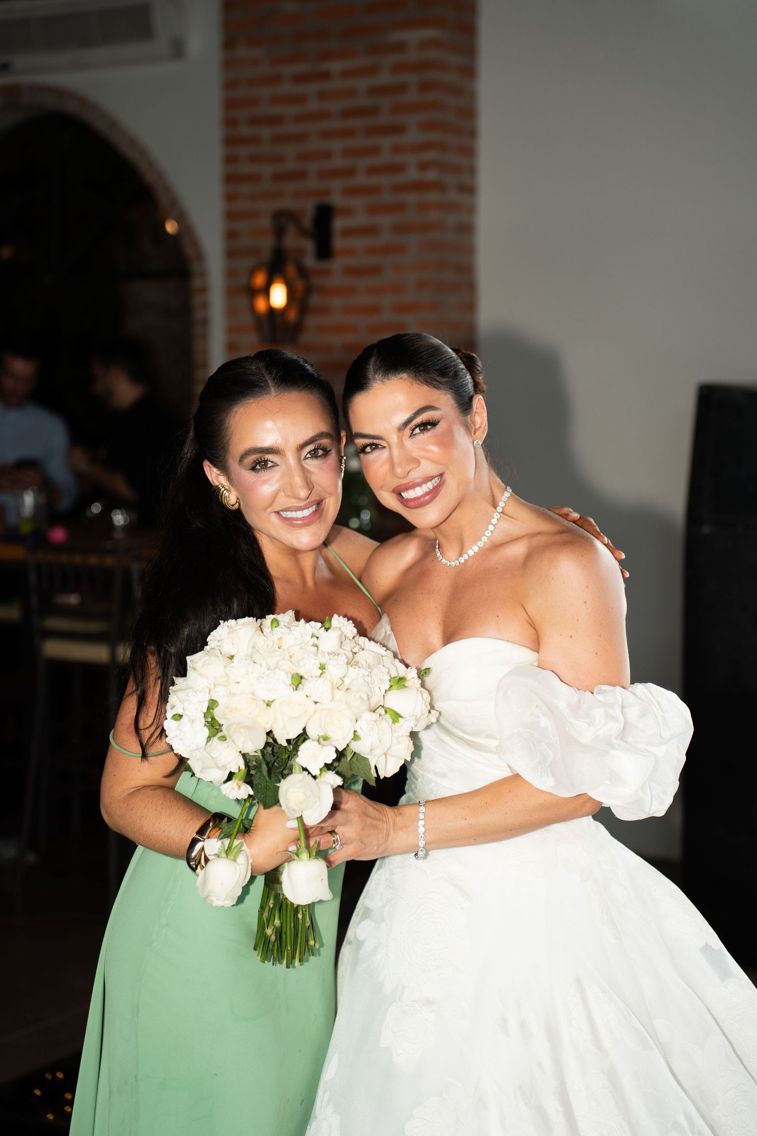 Bride in white gown holds white rose bouquet with woman in green dress at wedding reception.