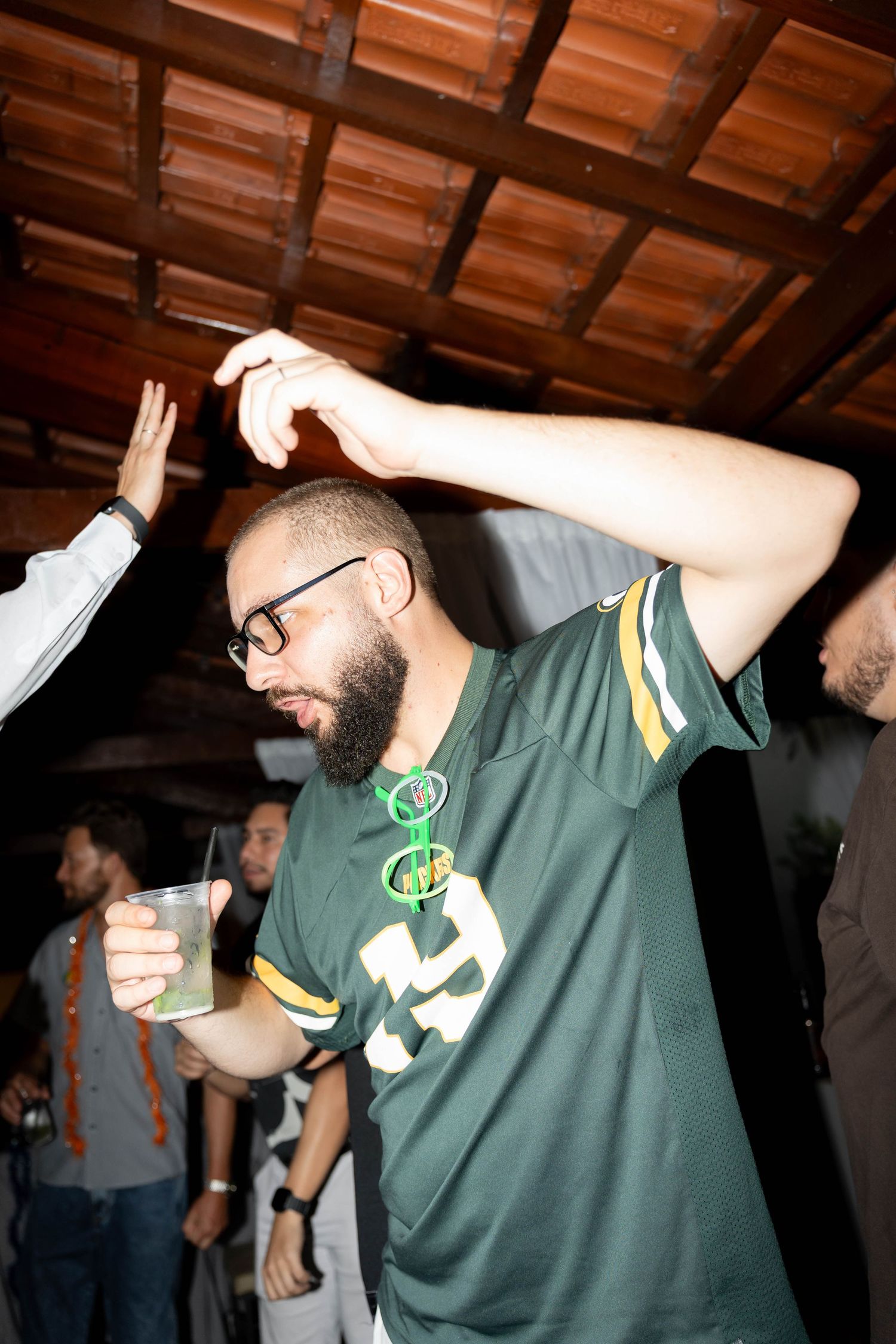 A bearded man in a Green Bay Packers jersey dances at a party while holding a drink.