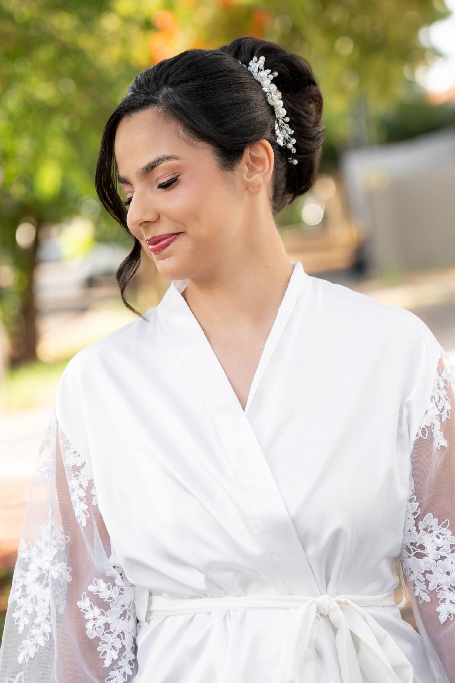 Bride wearing a white lace robe with floral hair accessory, smiling outdoors with green trees in the background.