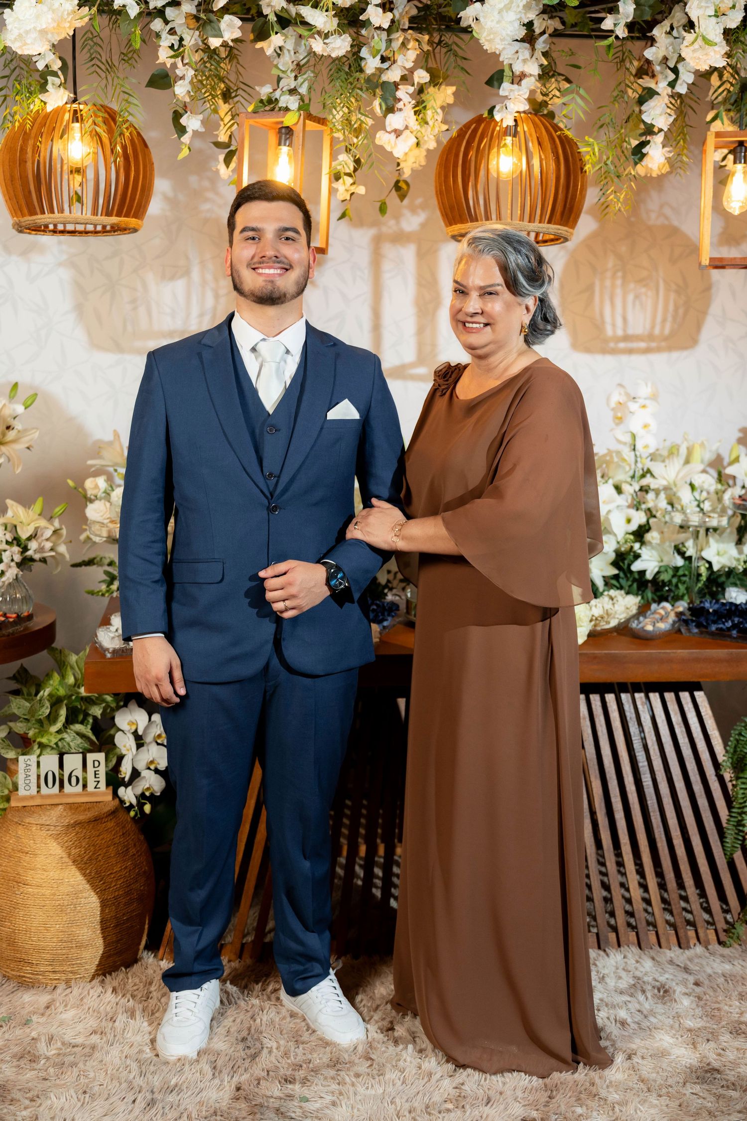 A young man in a navy blue suit poses with an older woman in a brown dress at an elegant floral event.