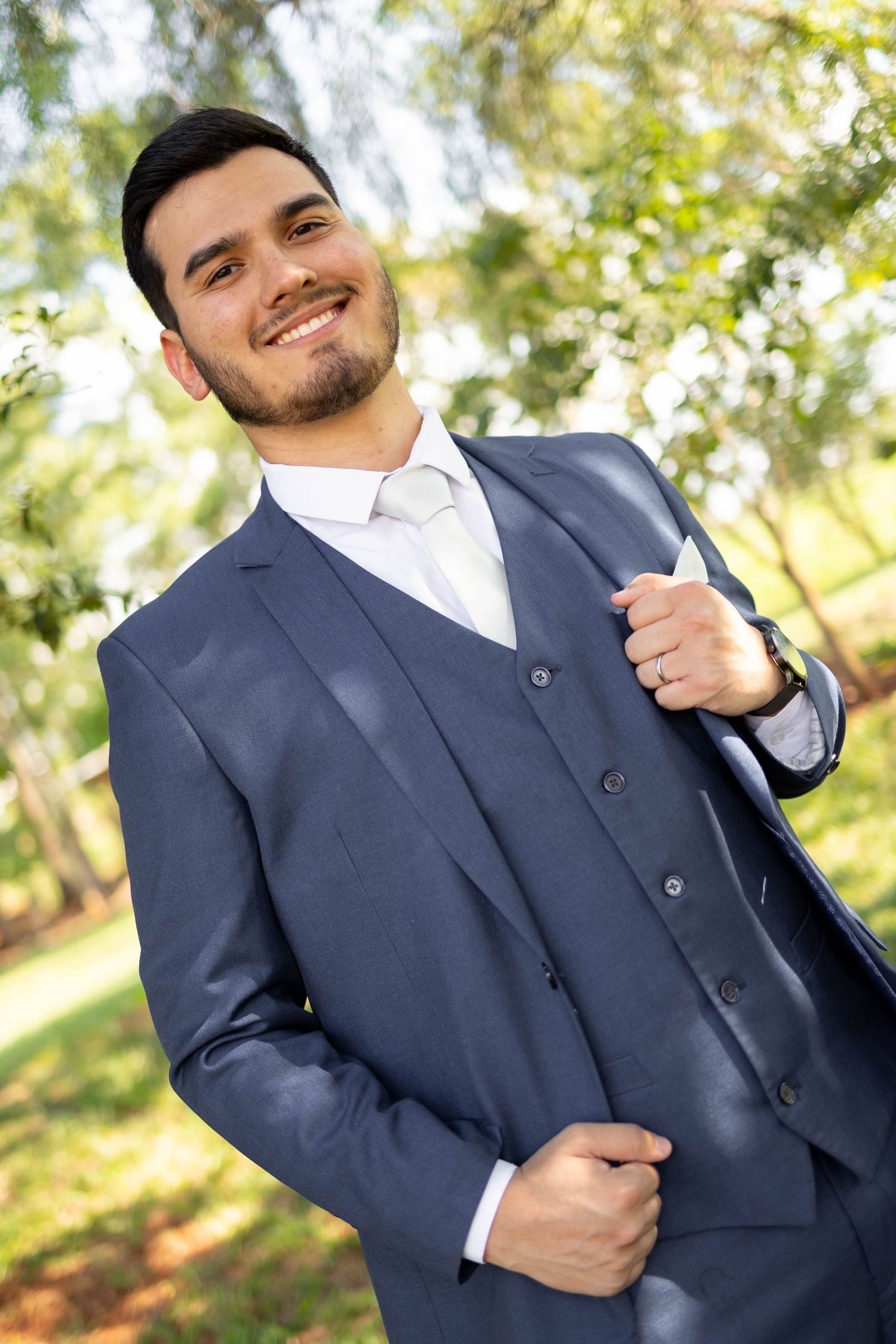 Smiling young man in navy blue three-piece suit with white tie posing outdoors in a park setting.