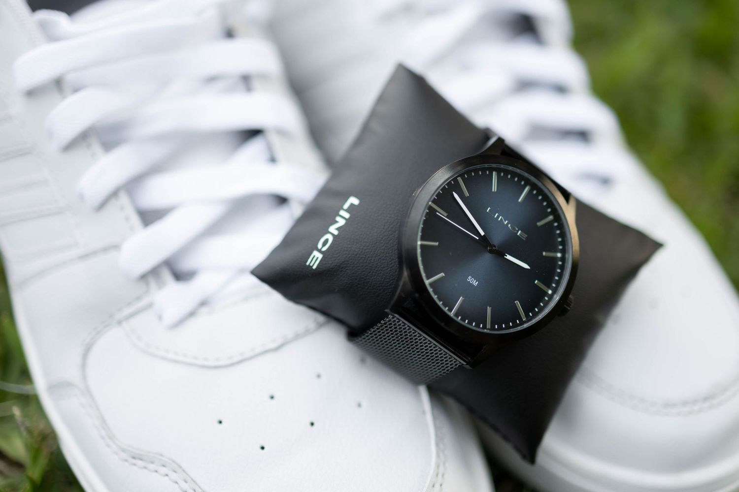 Black Lince minimalist watch resting on white Adidas sneakers outdoors on green grass.
