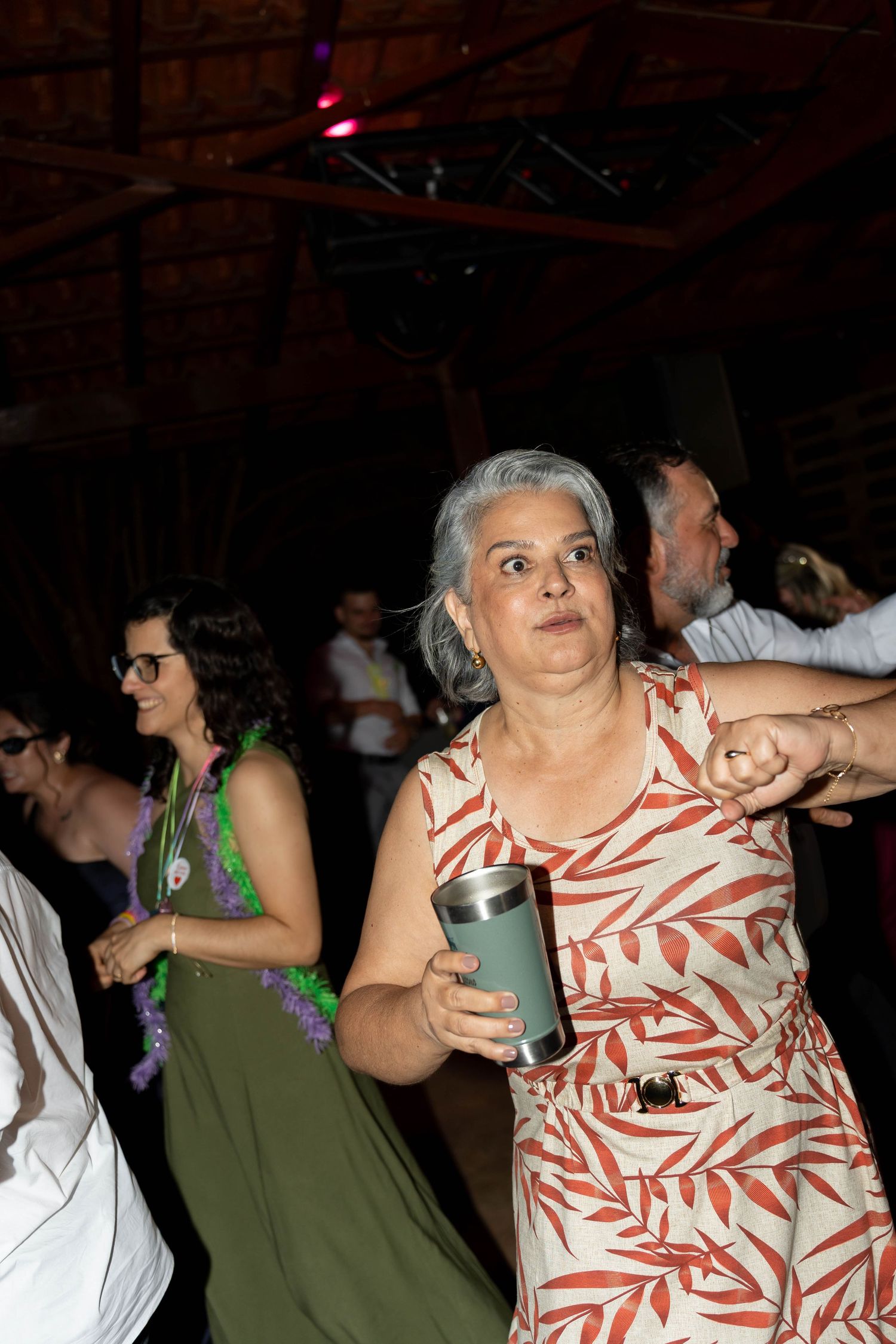 A gray-haired woman in a floral dress dances at a party, holding a tumbler cup, with guests celebrating in the background.