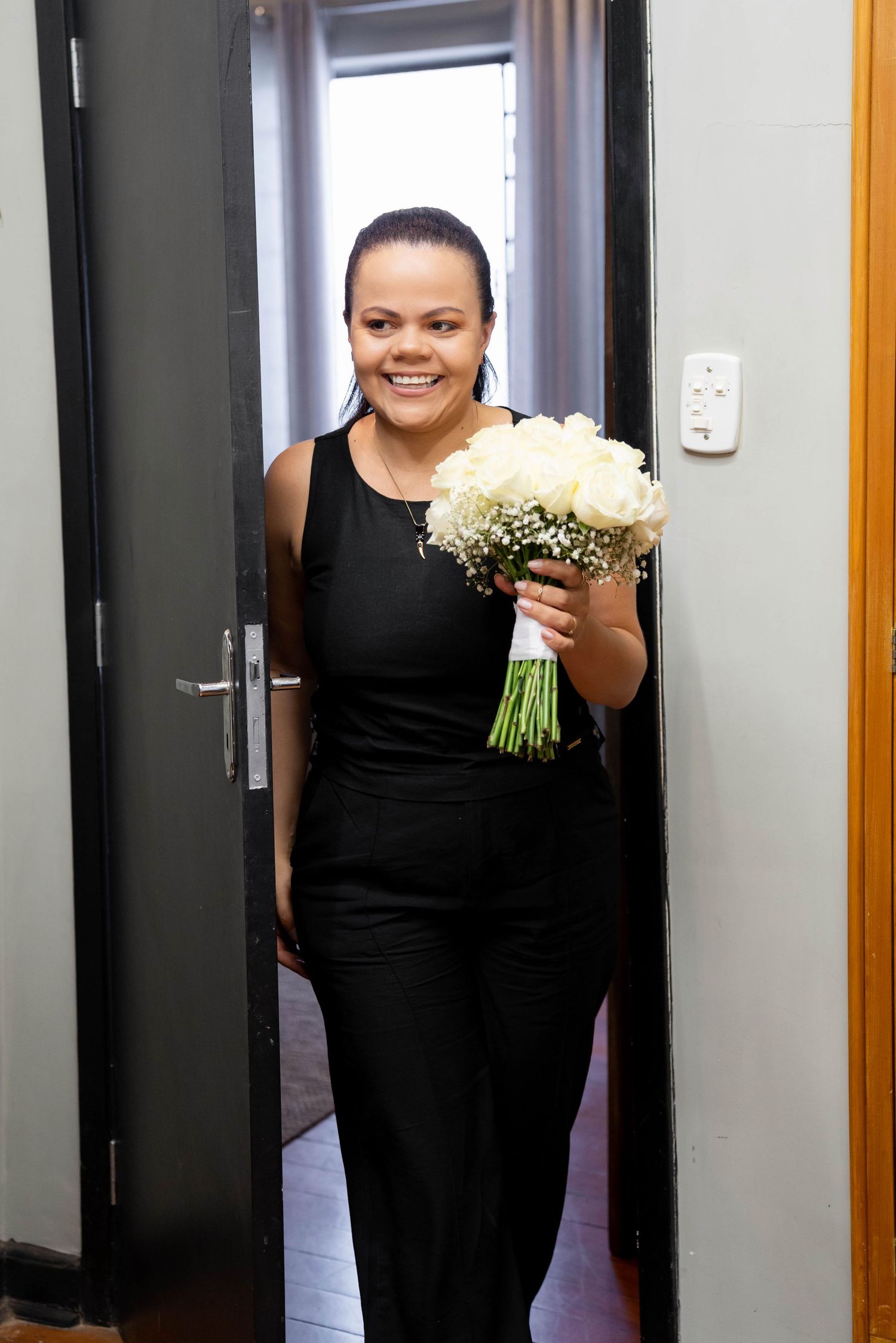 Smiling woman in black jumpsuit holding white rose bouquet standing in doorway.