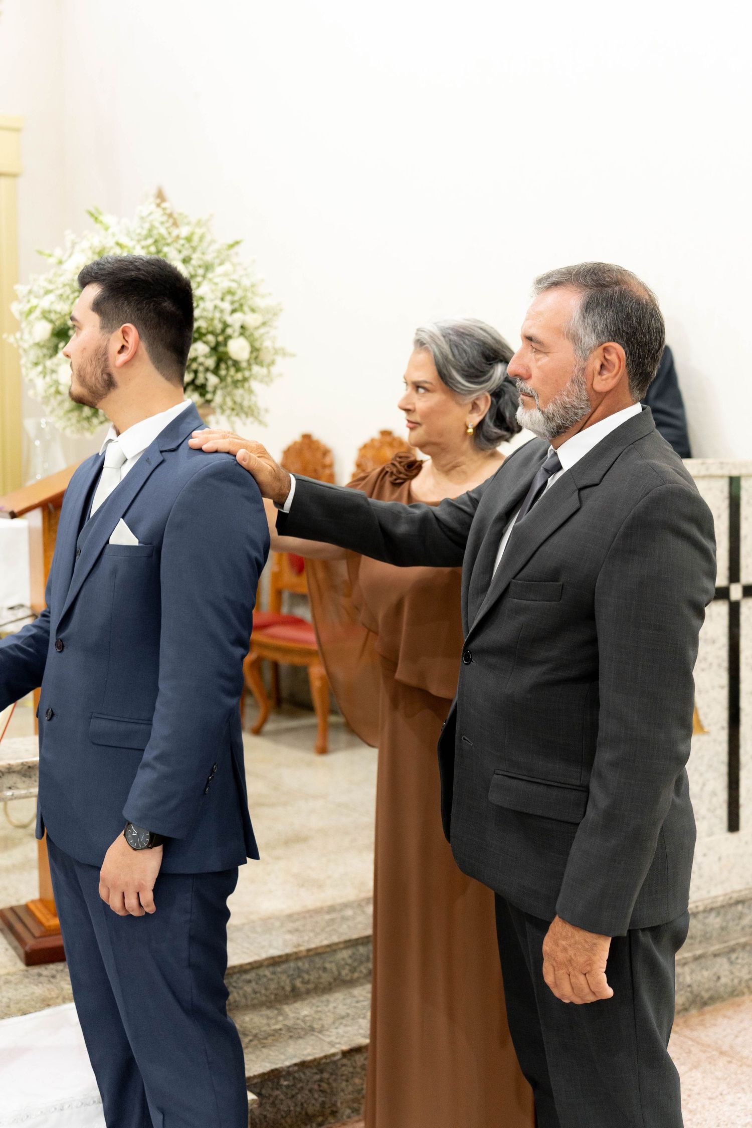 Groom in navy suit receives blessing from parents in church before wedding ceremony.