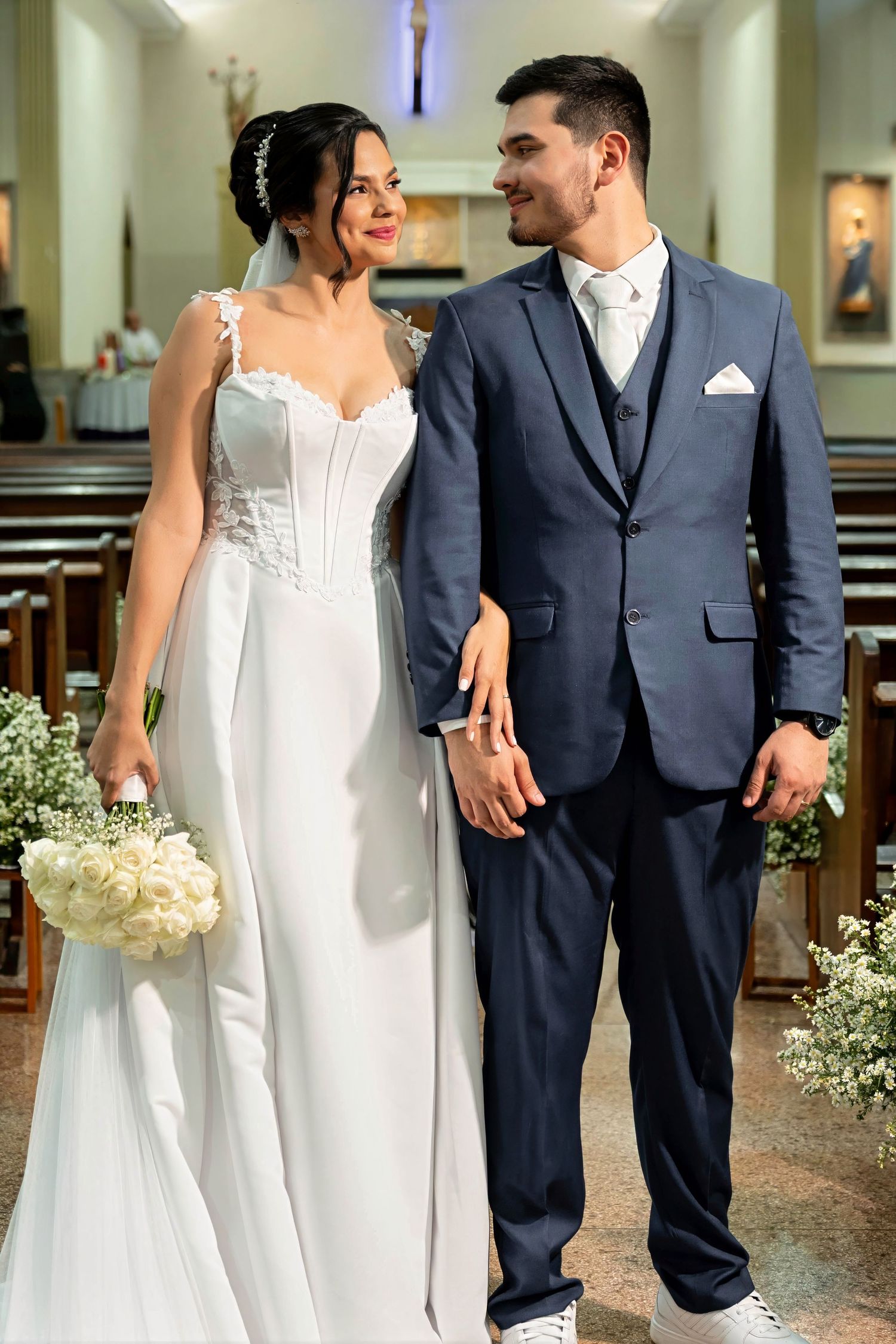 Bride in white gown holding bouquet and groom in navy suit walking together in church aisle.