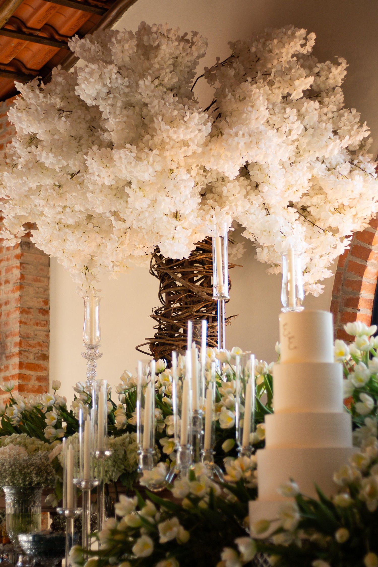 Elegant wedding reception with white floral trees, tall candles, and a tiered white cake surrounded by white flowers.
