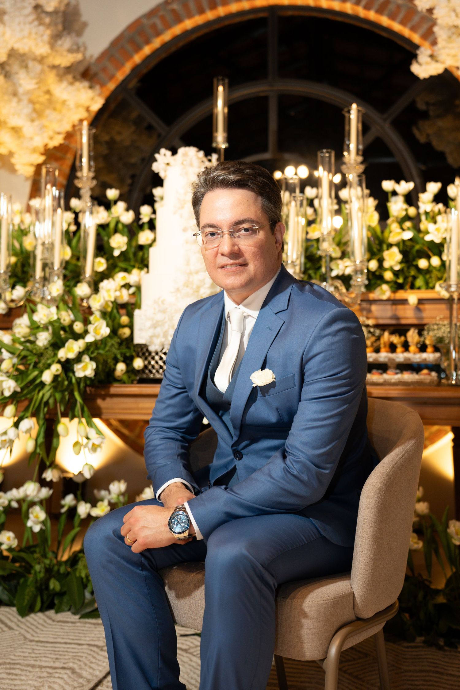 A man in a blue suit with white pocket square sits in a chair surrounded by elegant floral arrangements at a formal event.