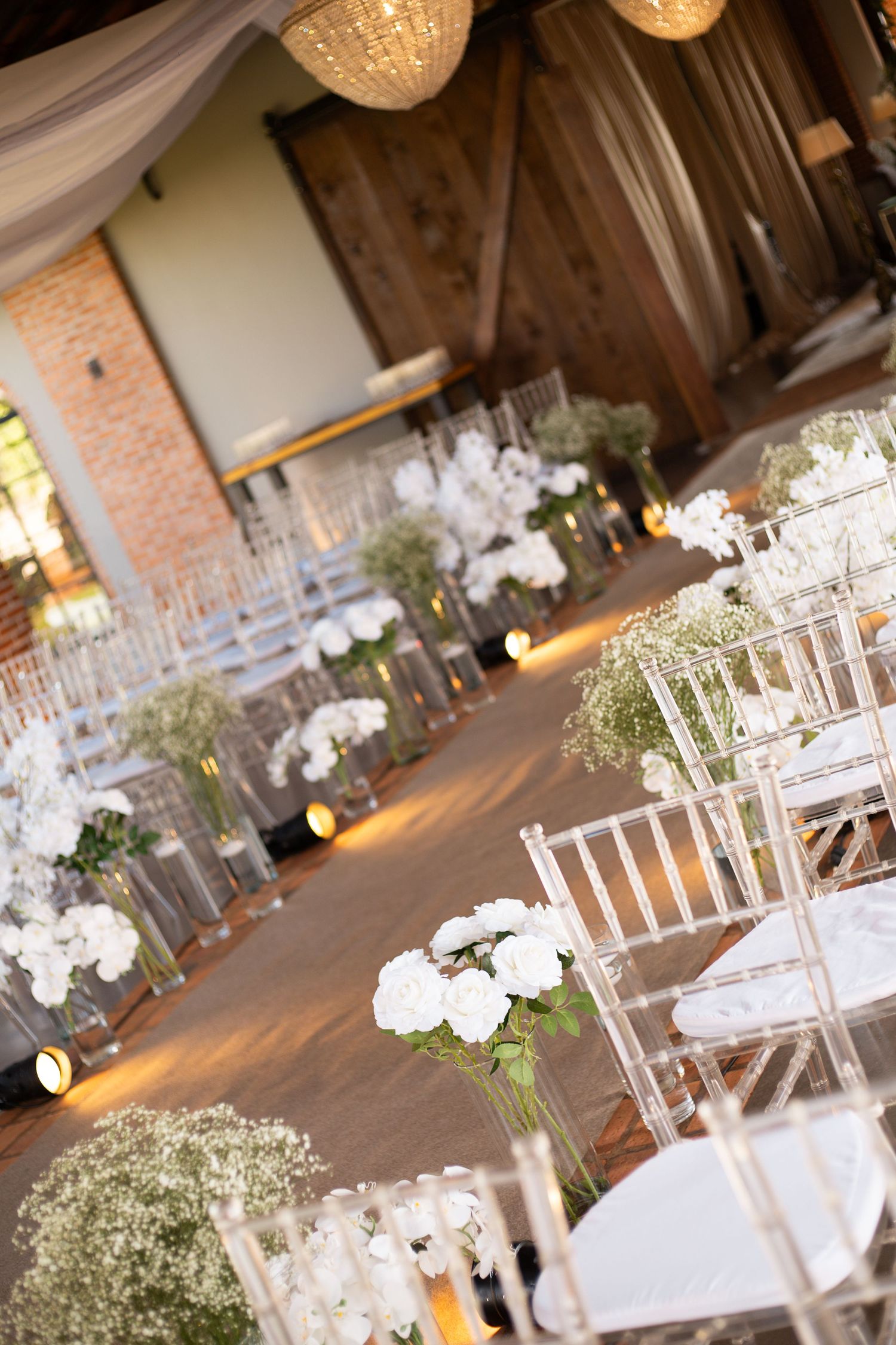 Elegant wedding ceremony aisle with white floral arrangements, baby's breath, and clear chiavari chairs in a rustic barn venue.