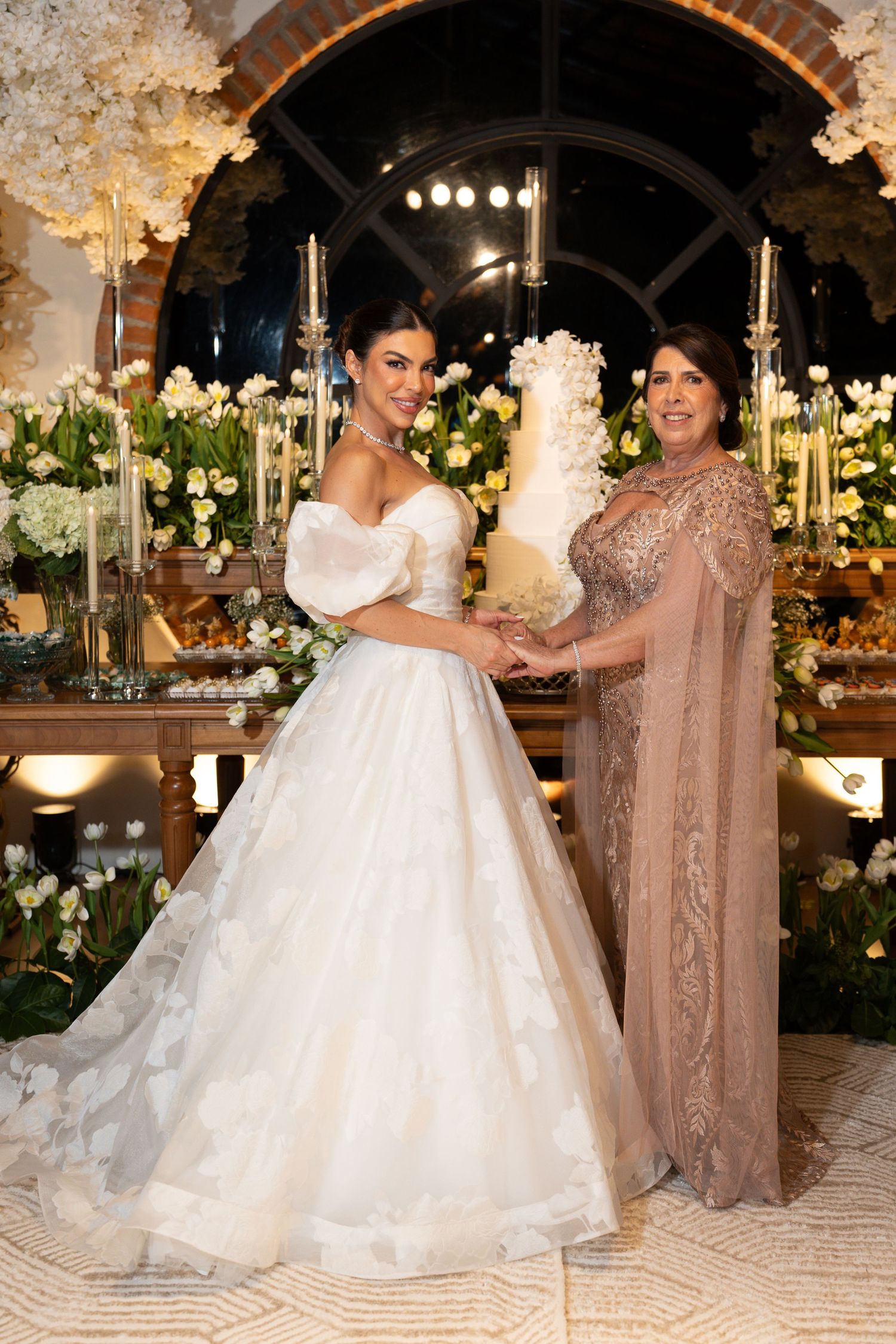 Bride in white ball gown and older woman in beige gown pose together near elegant wedding cake with floral decor.