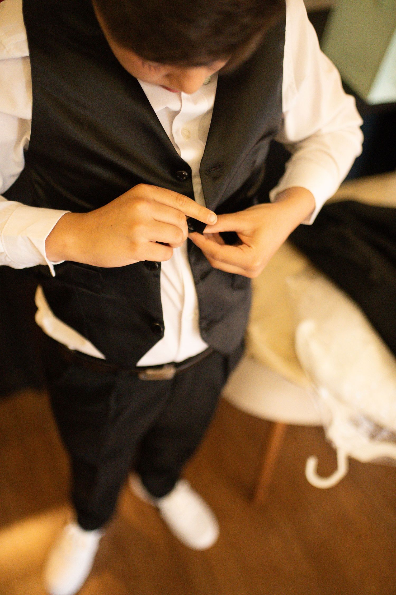 Young groom buttoning his black vest while getting dressed in a white shirt and dark trousers for a wedding ceremony.