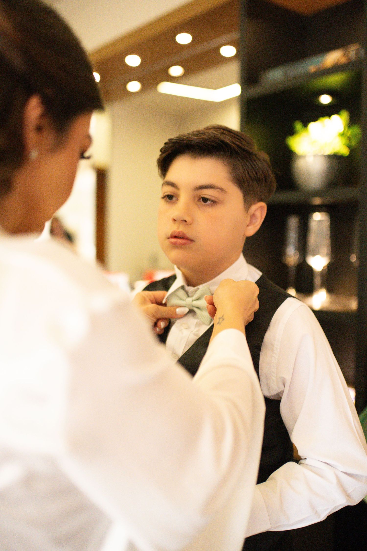 A woman adjusts a light blue bow tie on a young boy wearing a white shirt and black vest indoors.