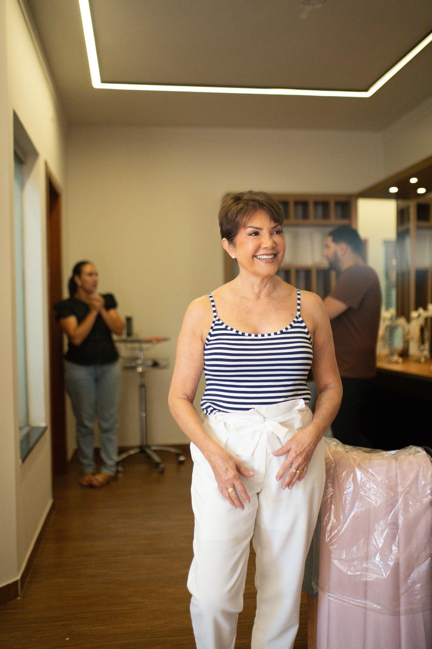 Smiling senior woman in striped top and white pants stands in a boutique fitting room with staff in background.