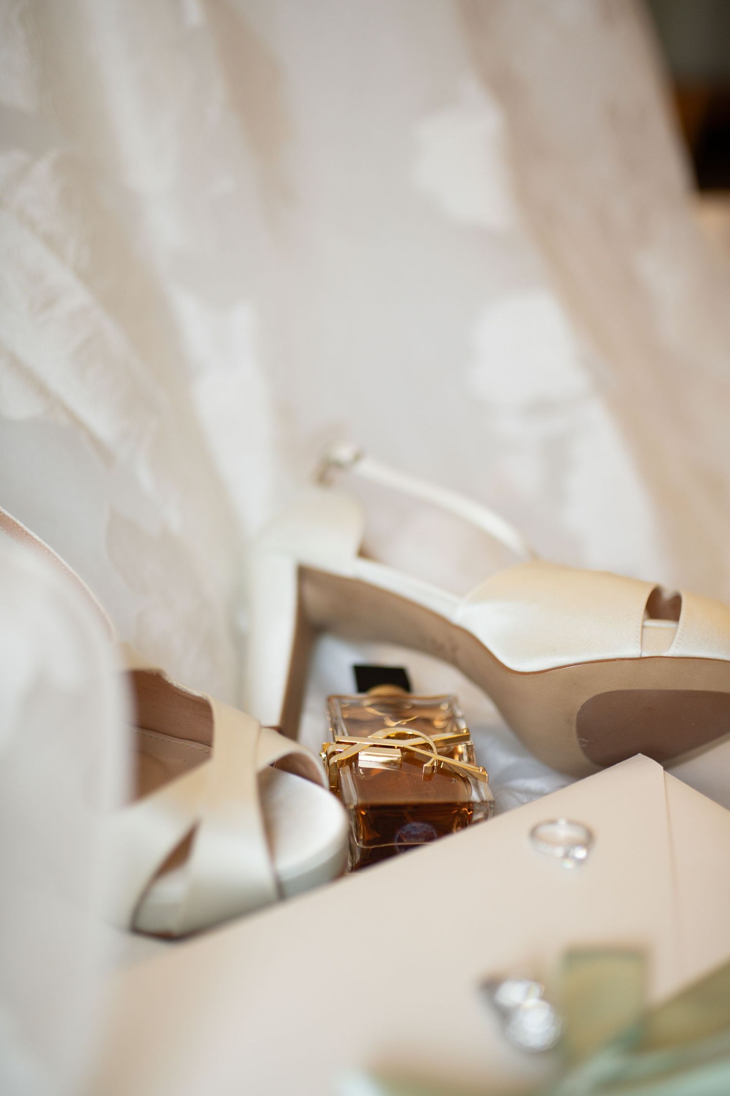 Elegant bridal accessories including ivory heels, YSL perfume, and diamond rings arranged beside a white wedding dress.