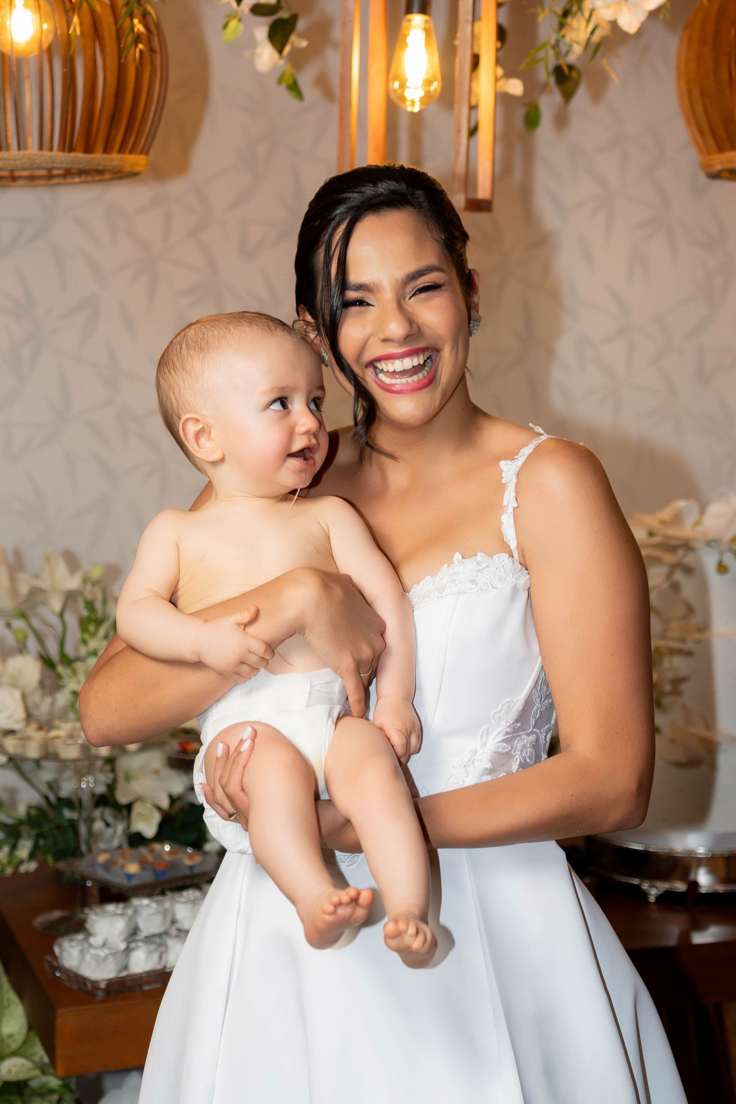 A joyful bride in a white wedding dress holds a smiling baby at a beautifully decorated wedding reception.