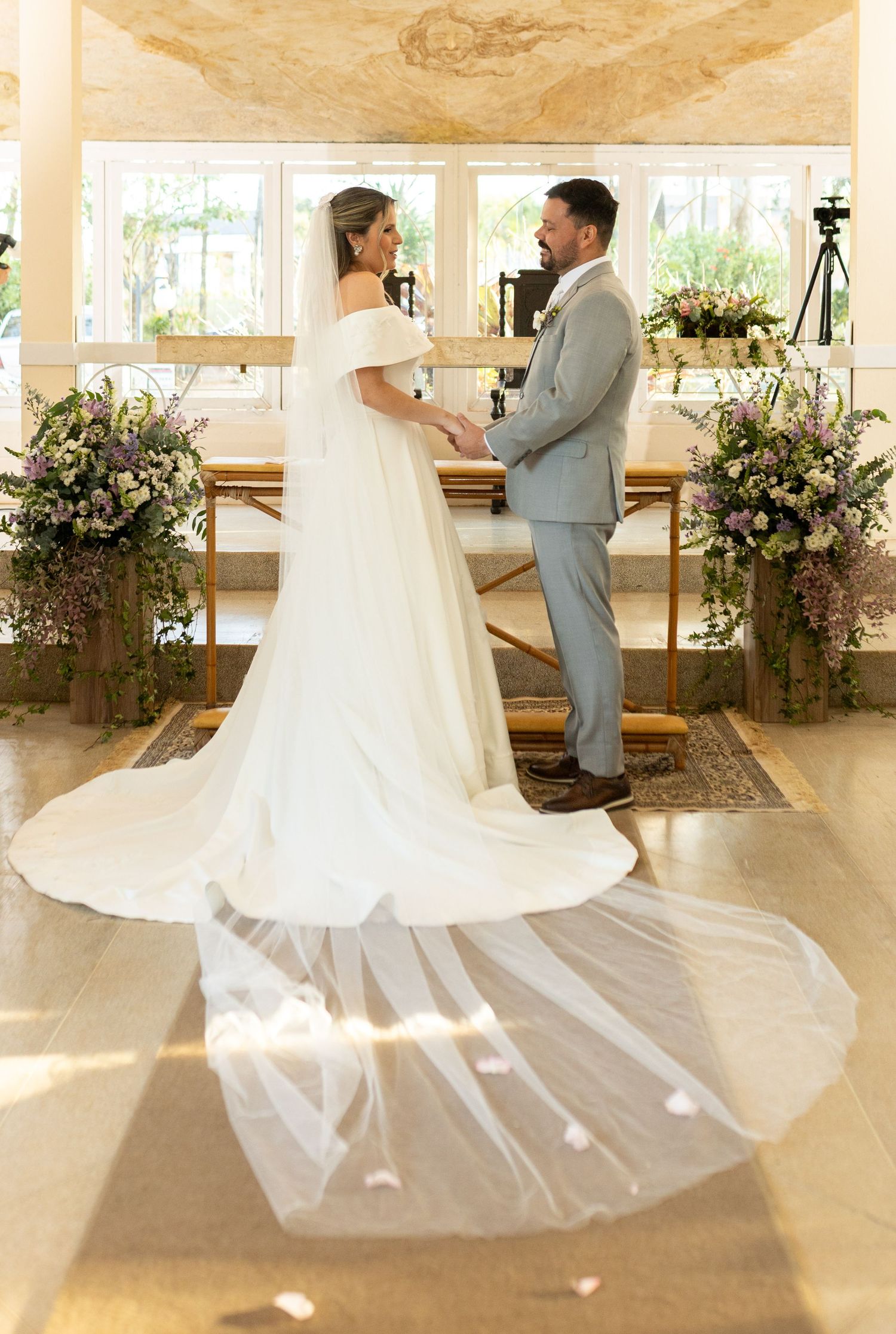 Bride and groom exchange vows at altar adorned with lush floral arrangements, bride wearing elegant off-shoulder gown with cathedral veil.