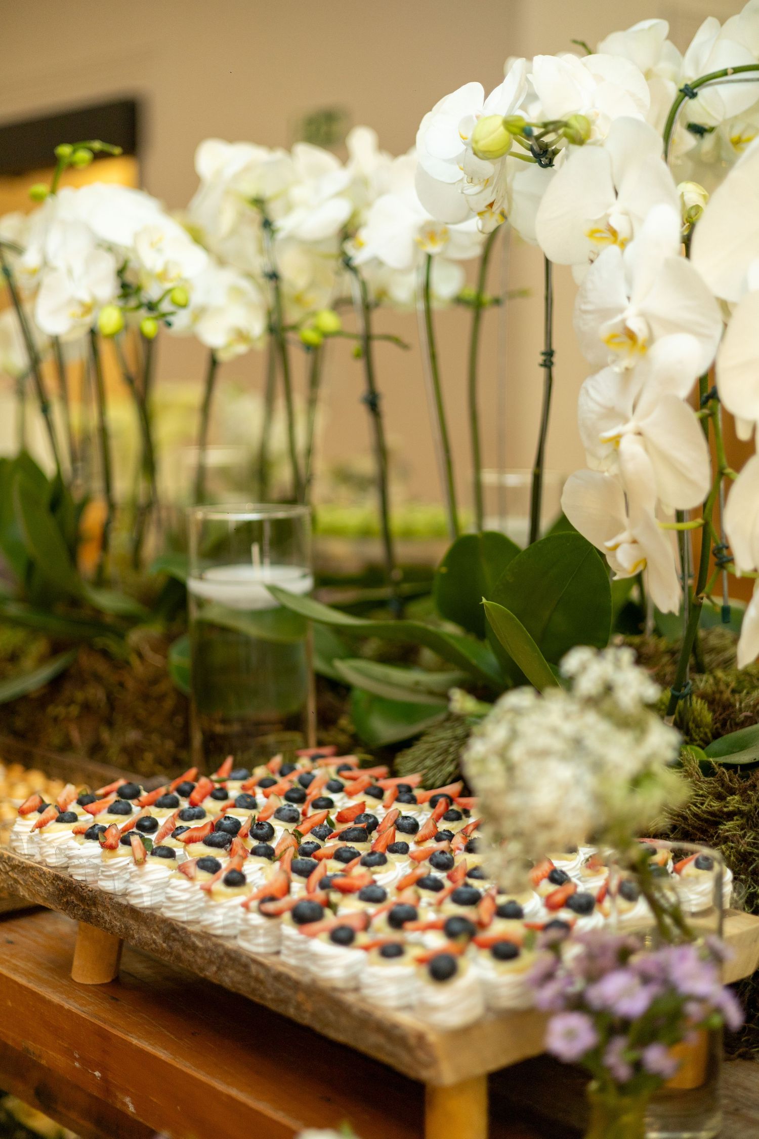 Elegant dessert display with berry-topped pastries on wooden board, surrounded by white orchids at an event.