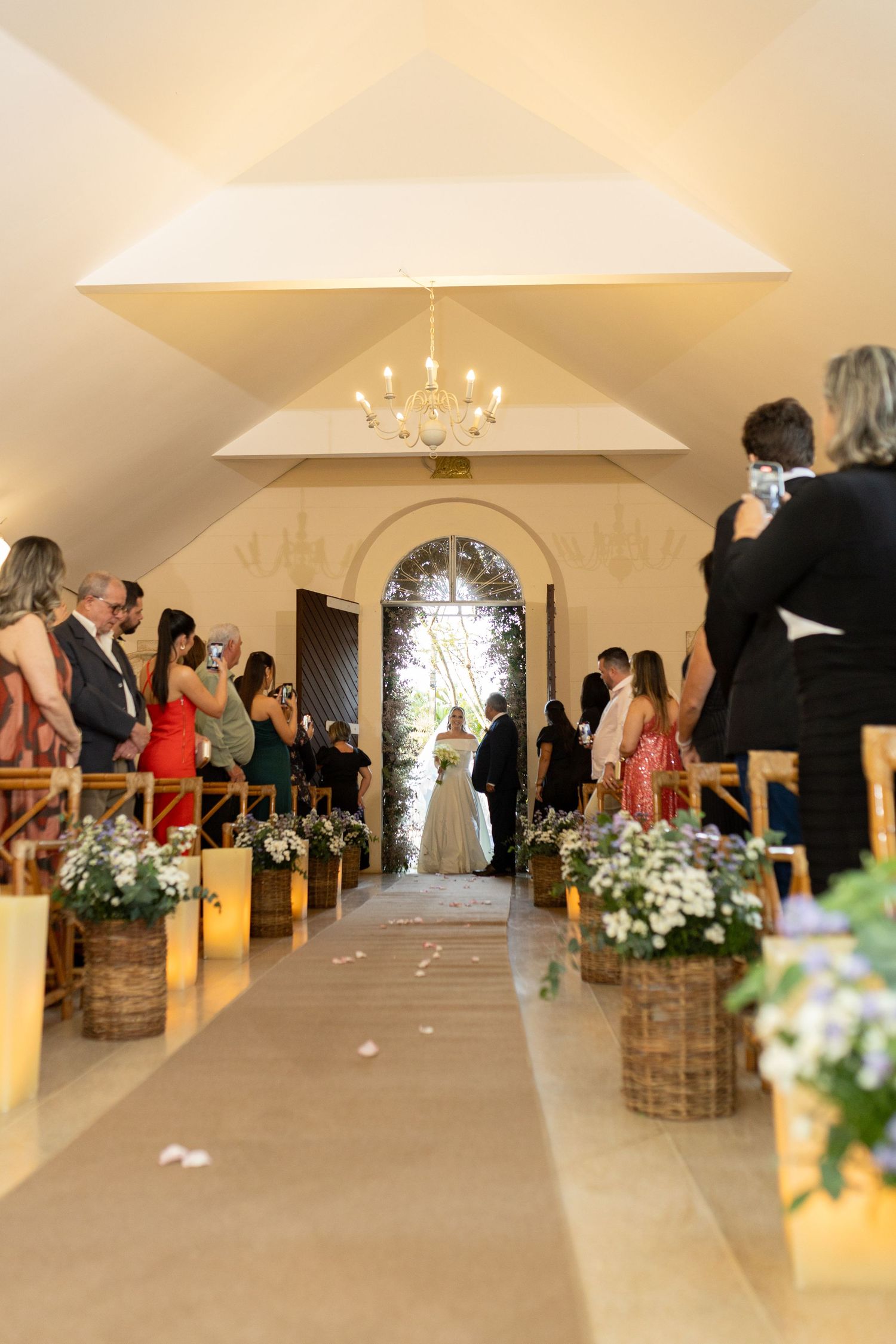 Bride walking down the aisle in an elegant chapel with candles and floral arrangements lining the path.