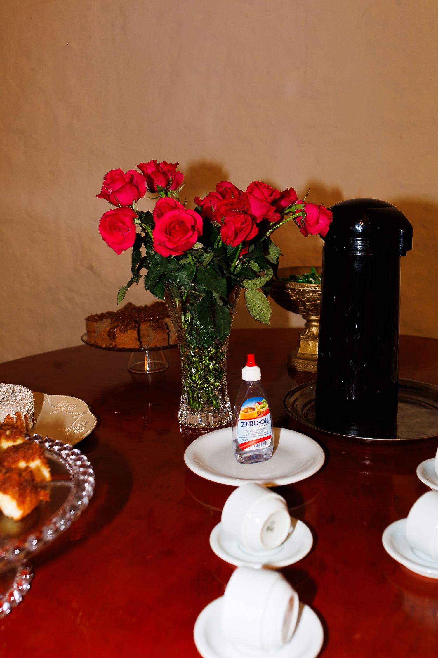 Red roses in glass vase on wooden table with white cups, black thermos, and Zero Cal sweetener at a tea setting.