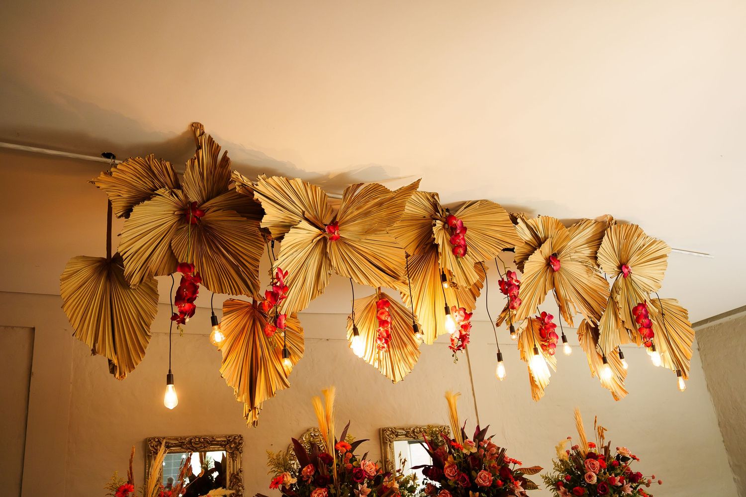Tropical dried palm leaf and red floral ceiling installation with warm Edison bulb lights for event decor.
