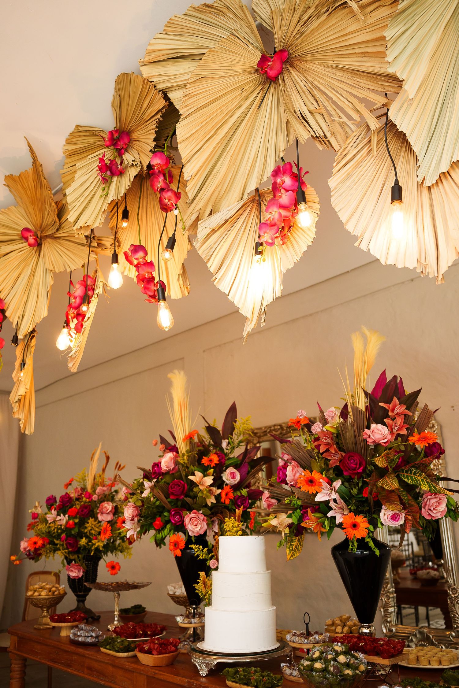 Elegant wedding dessert table with white tiered cake, colorful floral arrangements, and dried palm leaf ceiling decor.