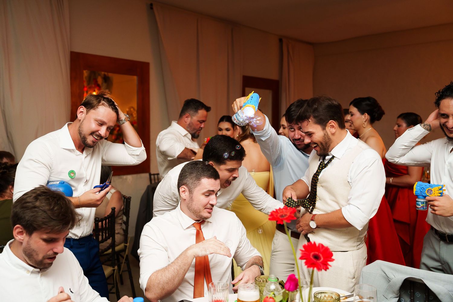 Lively wedding reception guests in white shirts celebrating and laughing together at decorated tables with colorful flowers.