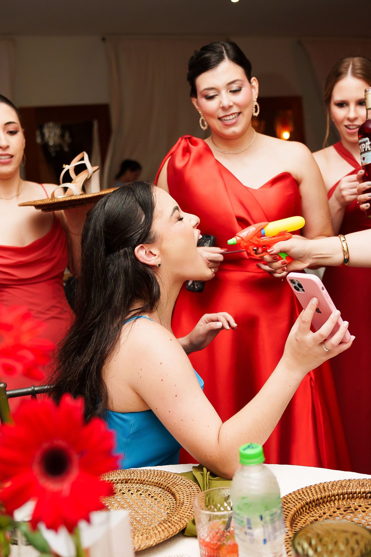 Women in red dresses at a party, one seated in blue taking a selfie while being offered colorful peppers.