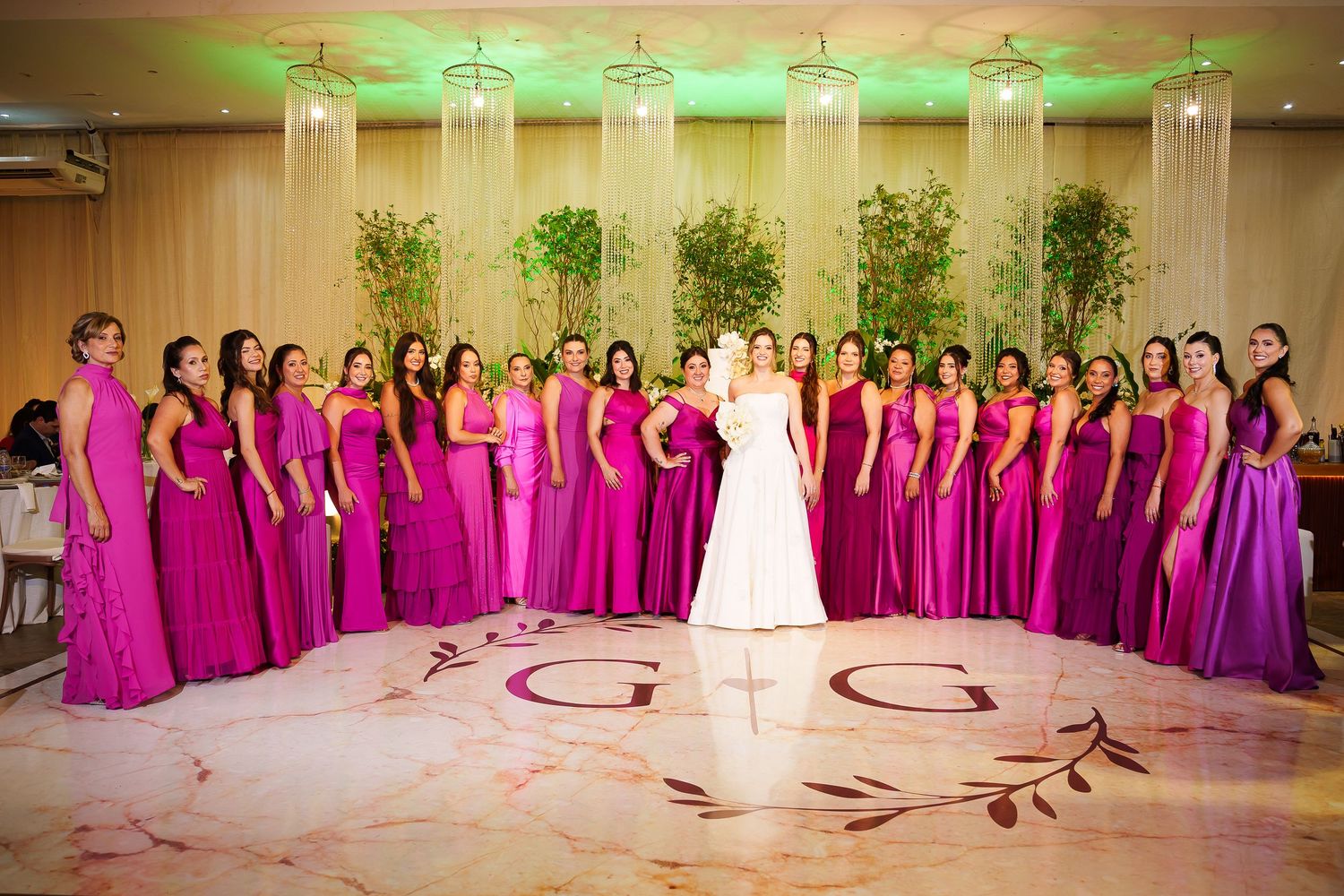 Bride in white gown surrounded by large bridal party in matching fuchsia gowns on monogrammed dance floor.