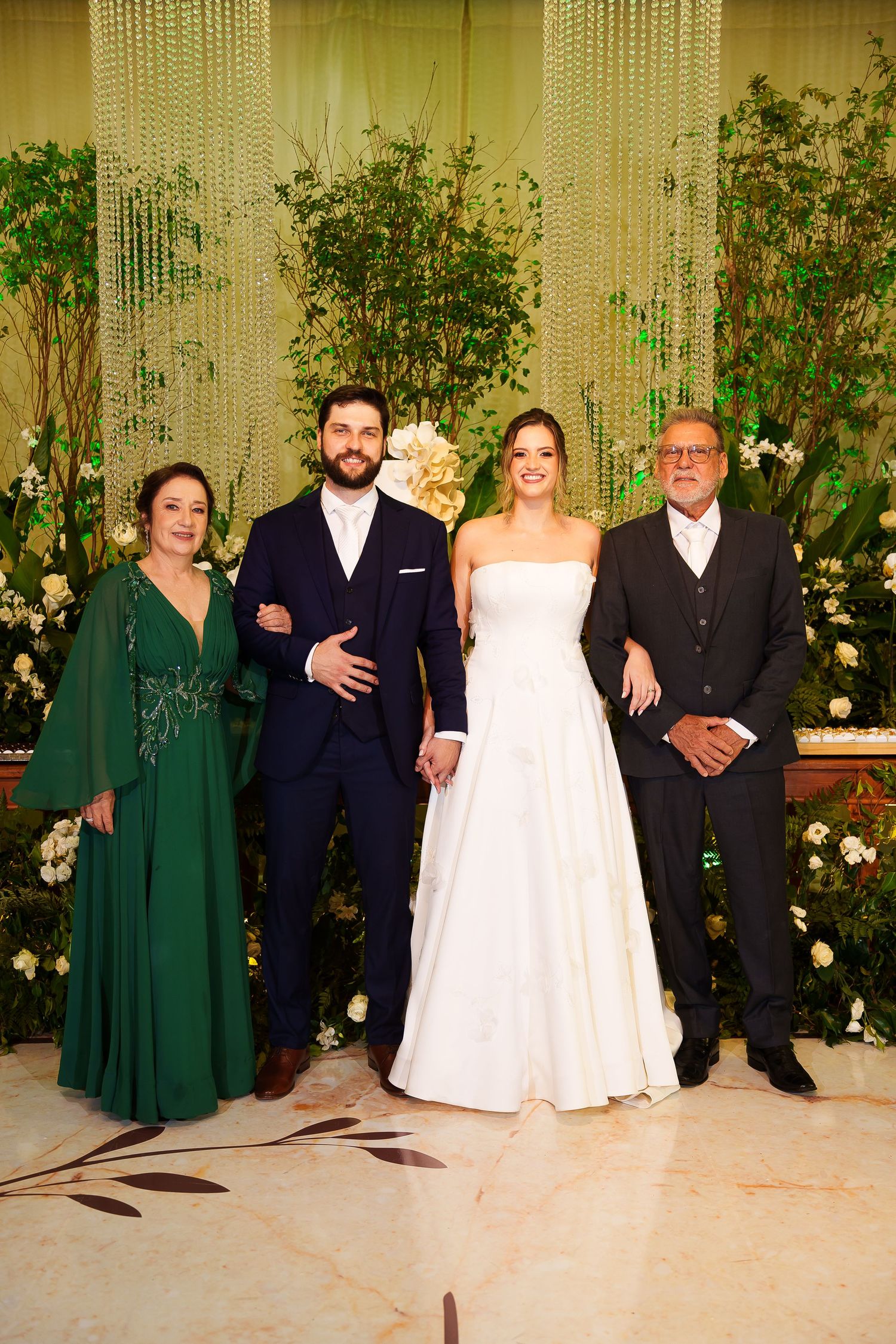 Wedding party of four poses elegantly before lush floral backdrop, bride in white gown flanked by groom and family.