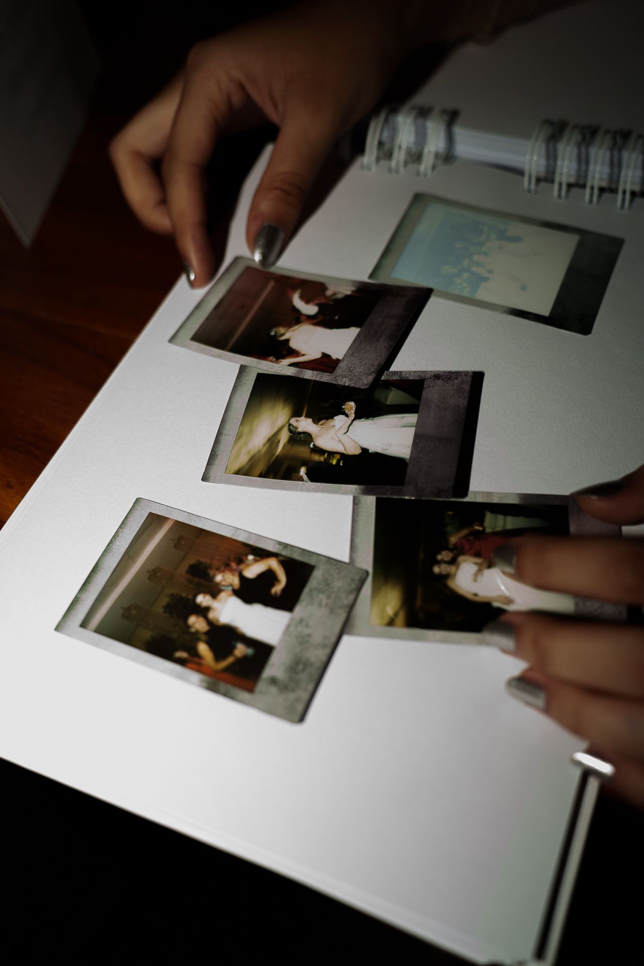 Hands with manicured nails arranging Polaroid photos in a spiral-bound photo album on a wooden table.