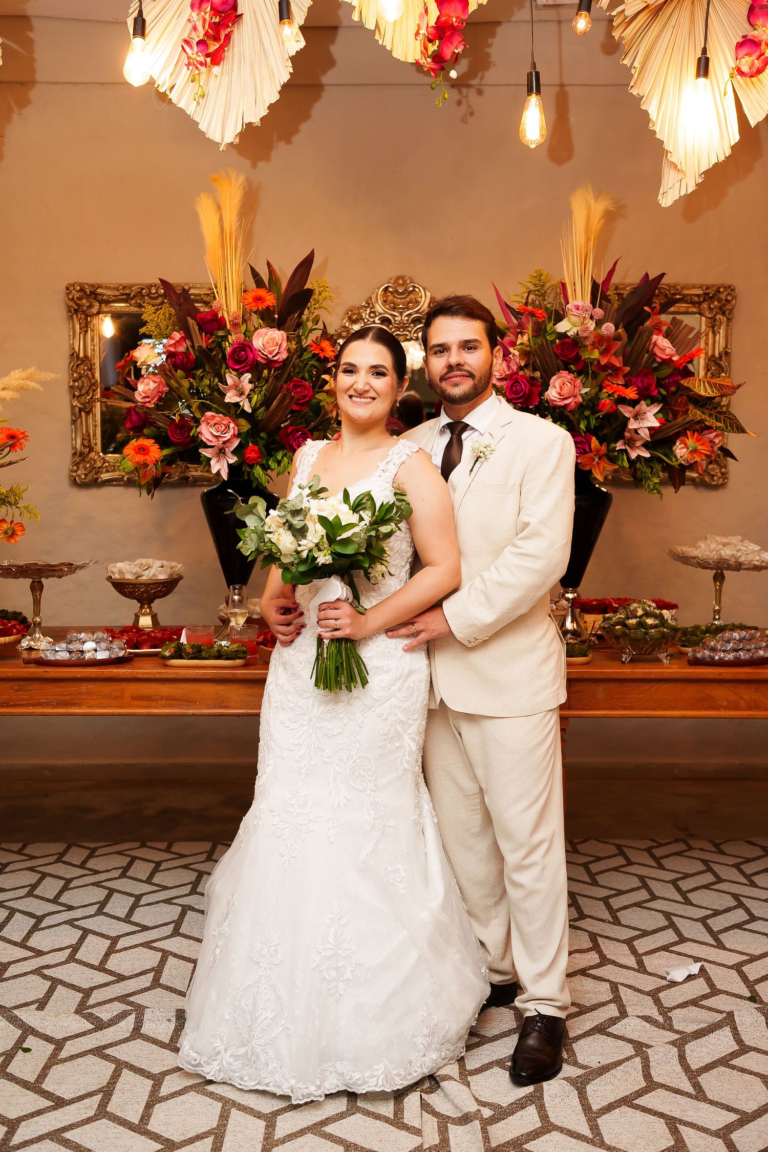 Bride in white lace gown holding bouquet stands with groom in cream suit before floral backdrop at wedding reception.
