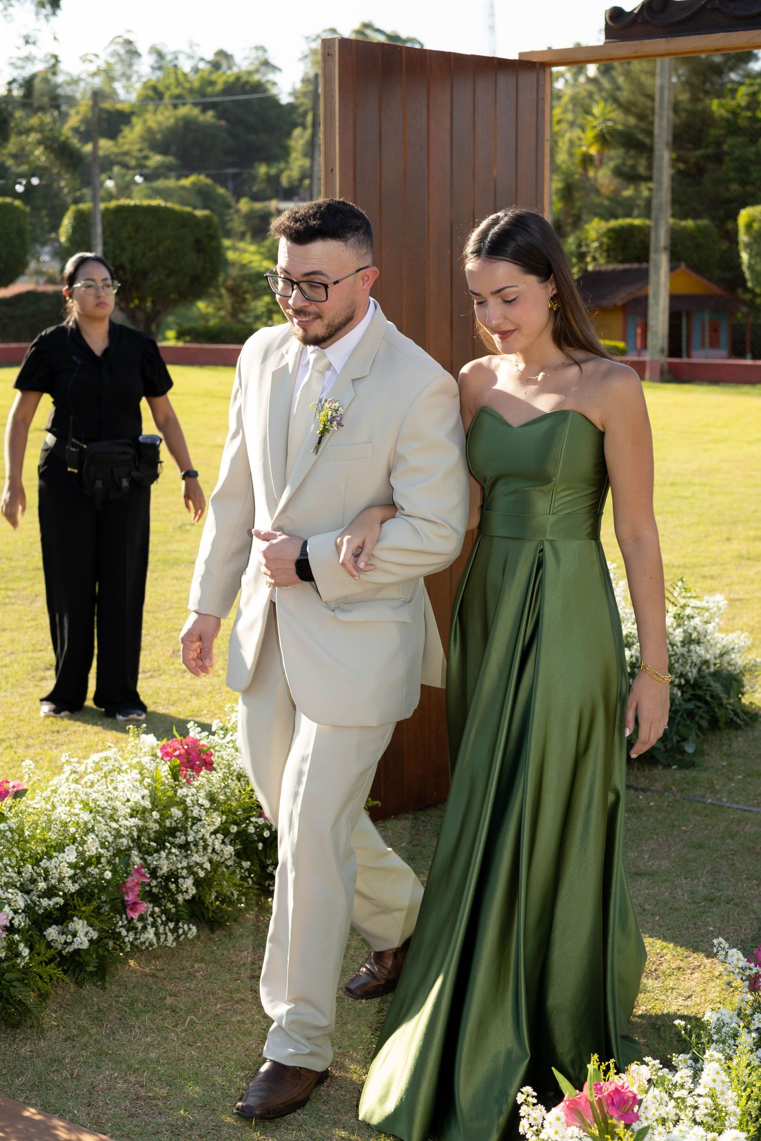 Groom in beige suit escorts bridesmaid in green satin gown at outdoor wedding ceremony with floral arrangements.